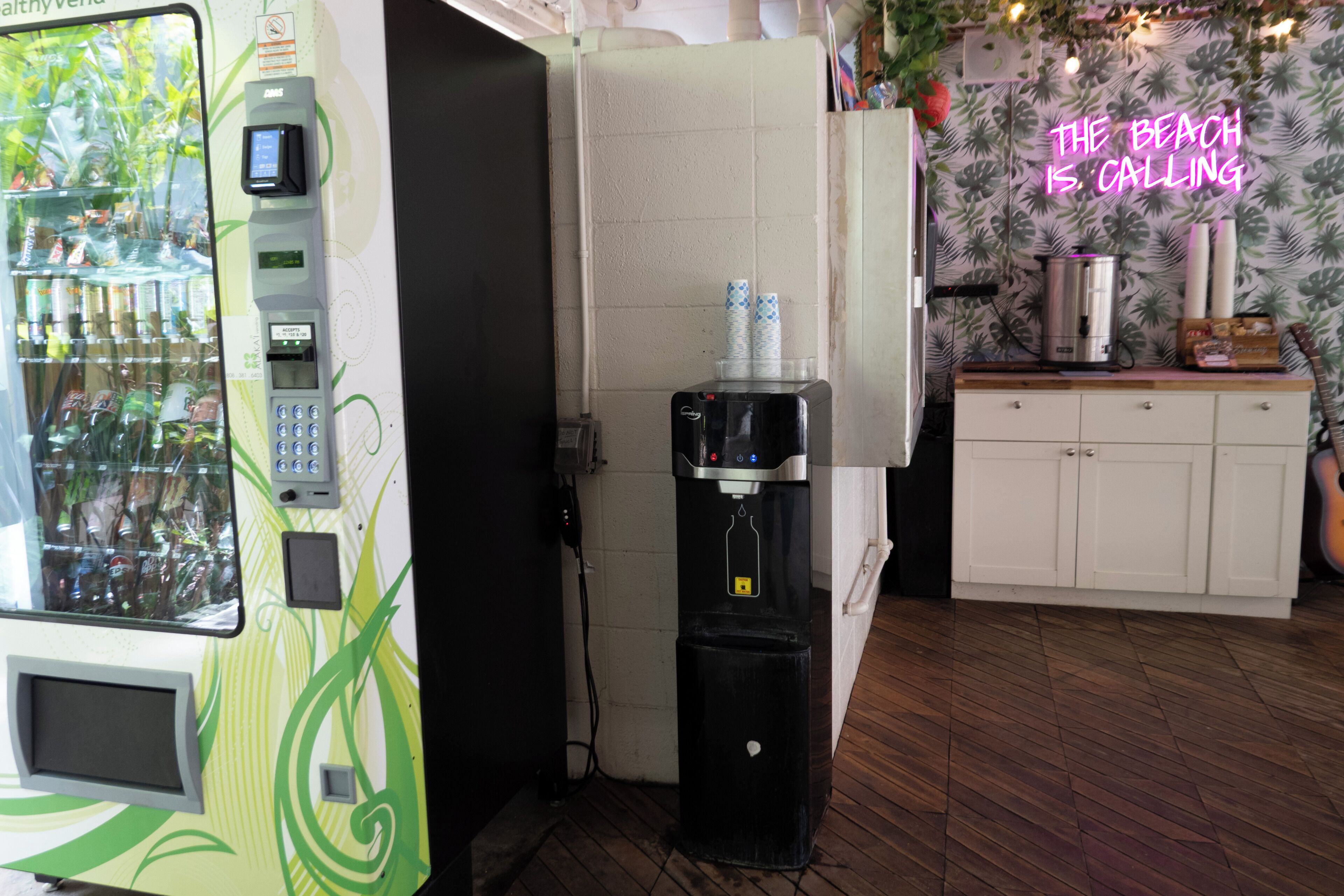 A vending machine, a water dispenser, and a neon sign in a room with wooden flooring and a white cabinet.