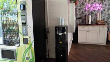 A vending machine, a water dispenser, and a neon sign in a room with wooden flooring and a white cabinet.