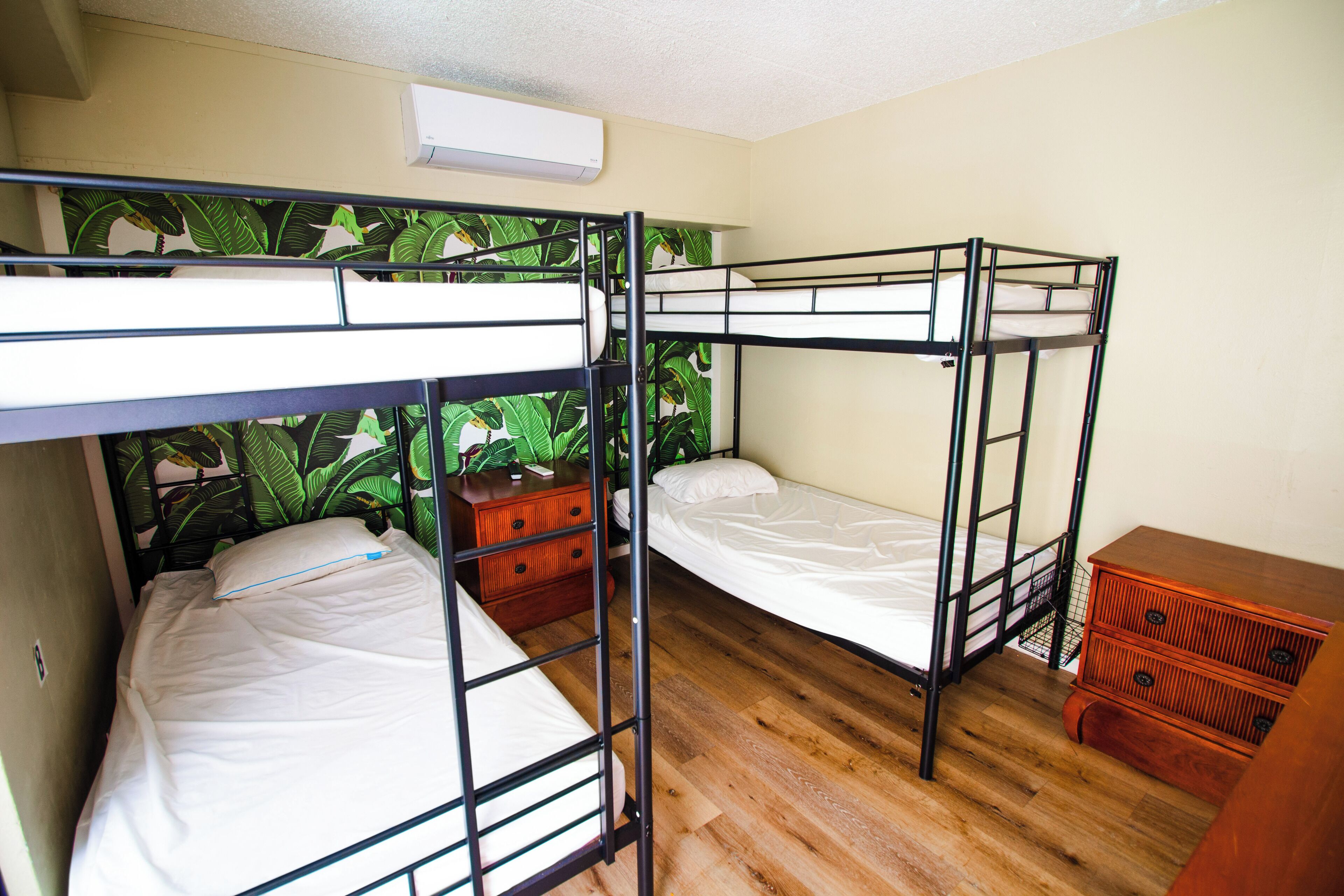 A bunk bed room with a wooden floor, a small dresser, and a wall with a tropical wallpaper.