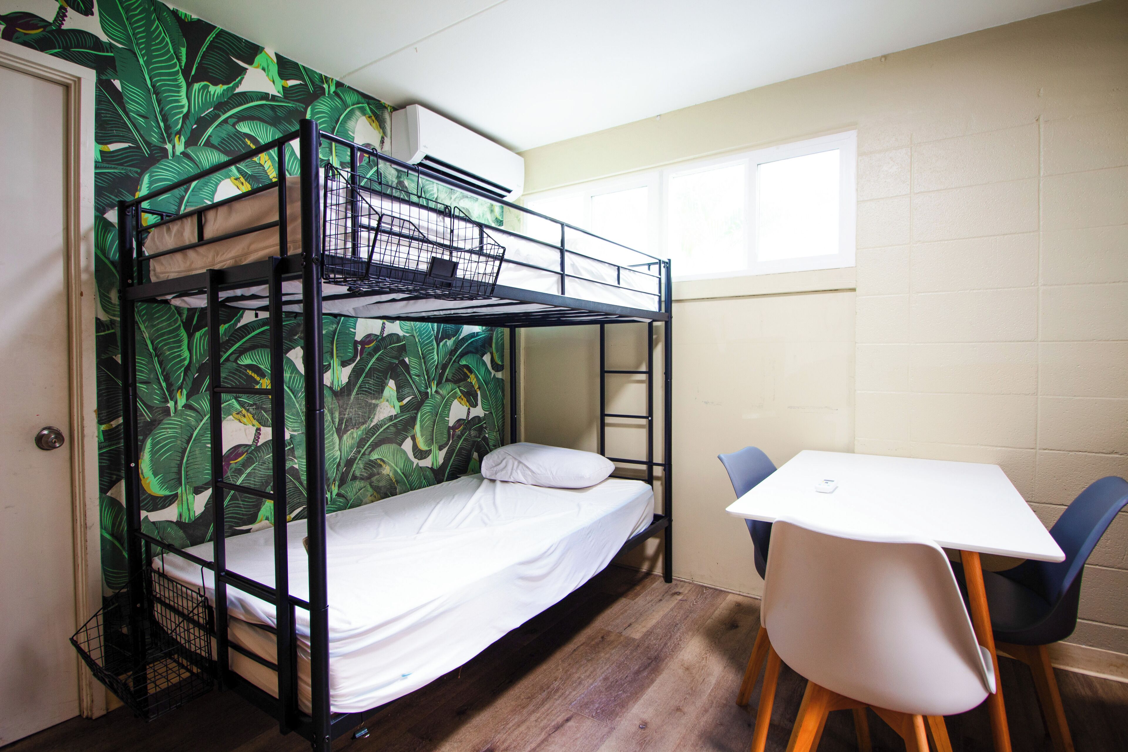 A bunk bed with a white mattress and a black metal frame, a white table with two chairs, and a wall with a green leaf pattern.