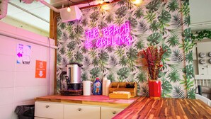 Eine Küche mit einer Neonreklame mit der Aufschrift 'THE BEACH IS CALLING', einer Kaffeemaschine, einem Mülleimer und einer Holzarbeitsplatte.