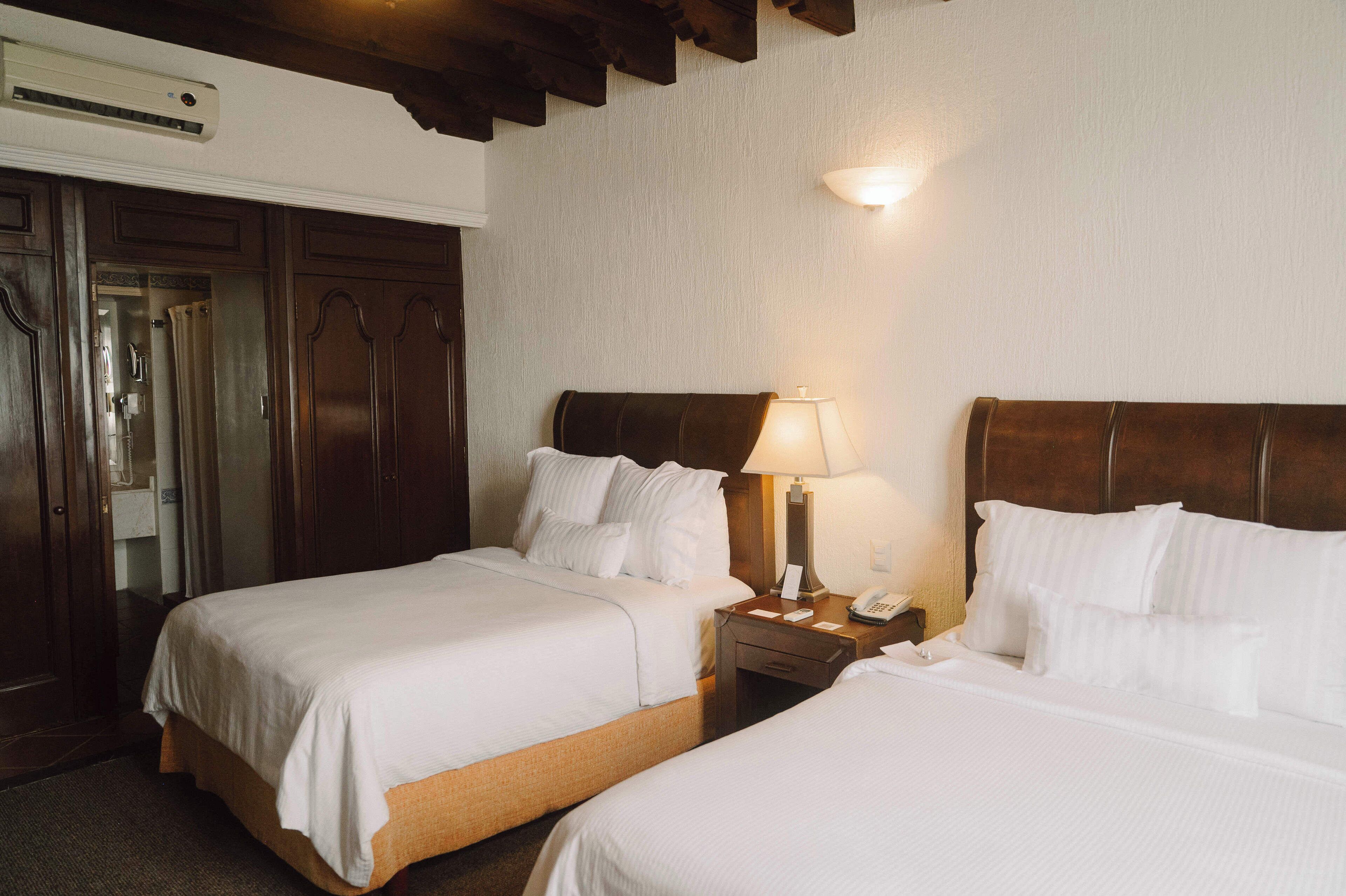 A hotel room with two beds, wooden headboards, a nightstand with a lamp, and a wall-mounted air conditioner.