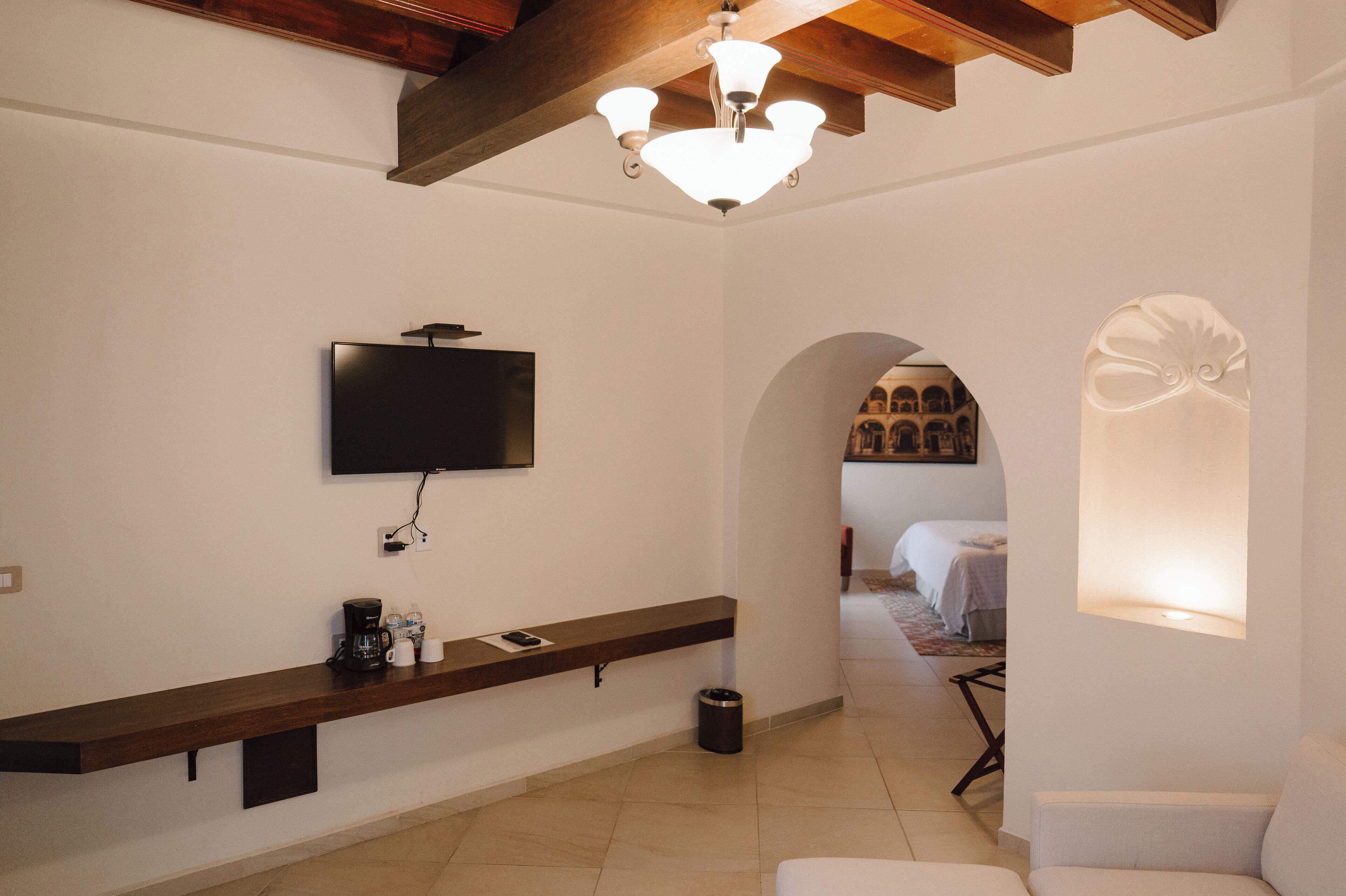Quarto de hotel moderno com televisão de tela plana fixada na parede, um banco de madeira com cafeteira, um cesto de lixo e uma poltrona branca.