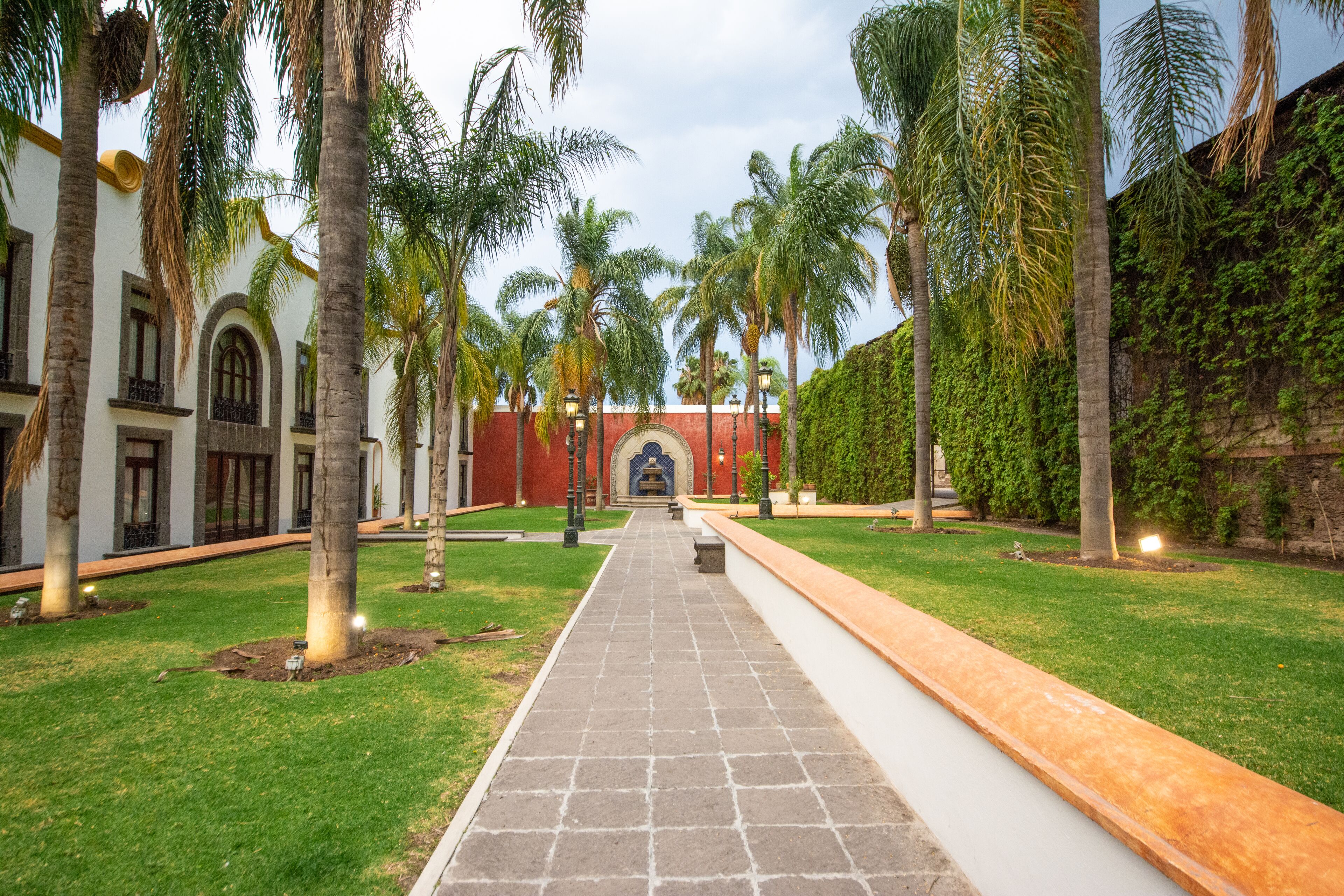 A courtyard with a paved walkway, palm trees, and a building with arched windows and a red wall.
