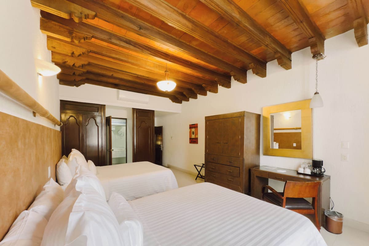 Une chambre d’hôtel avec deux lits, des poutres apparentes en bois au plafond, un bureau avec un miroir et une petite poubelle.