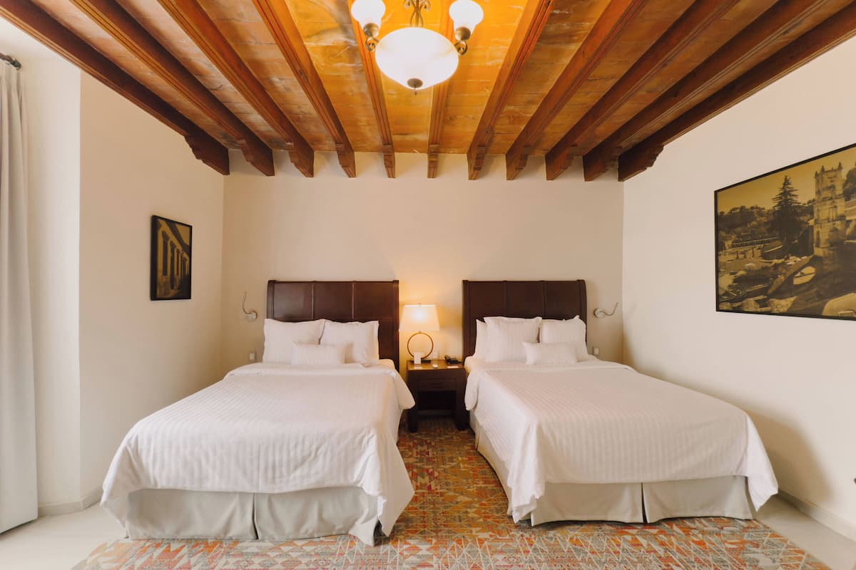 Une chambre d’hôtel avec deux lits, des poutres en bois au plafond, un sol à motifs et un tableau au mur.