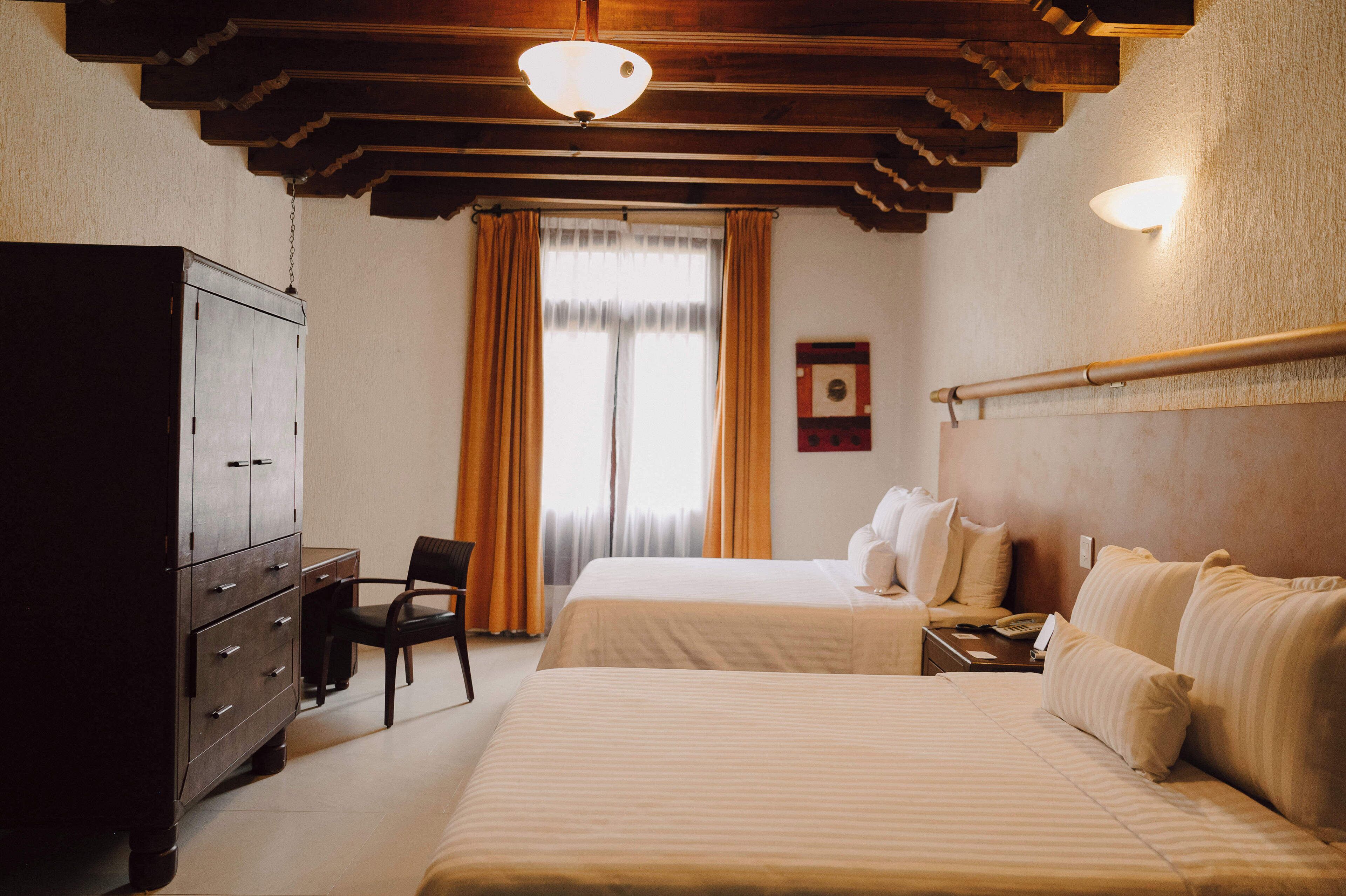 Habitación de hotel con dos camas, un aparador de madera, una silla y un ventanal con cortinas.