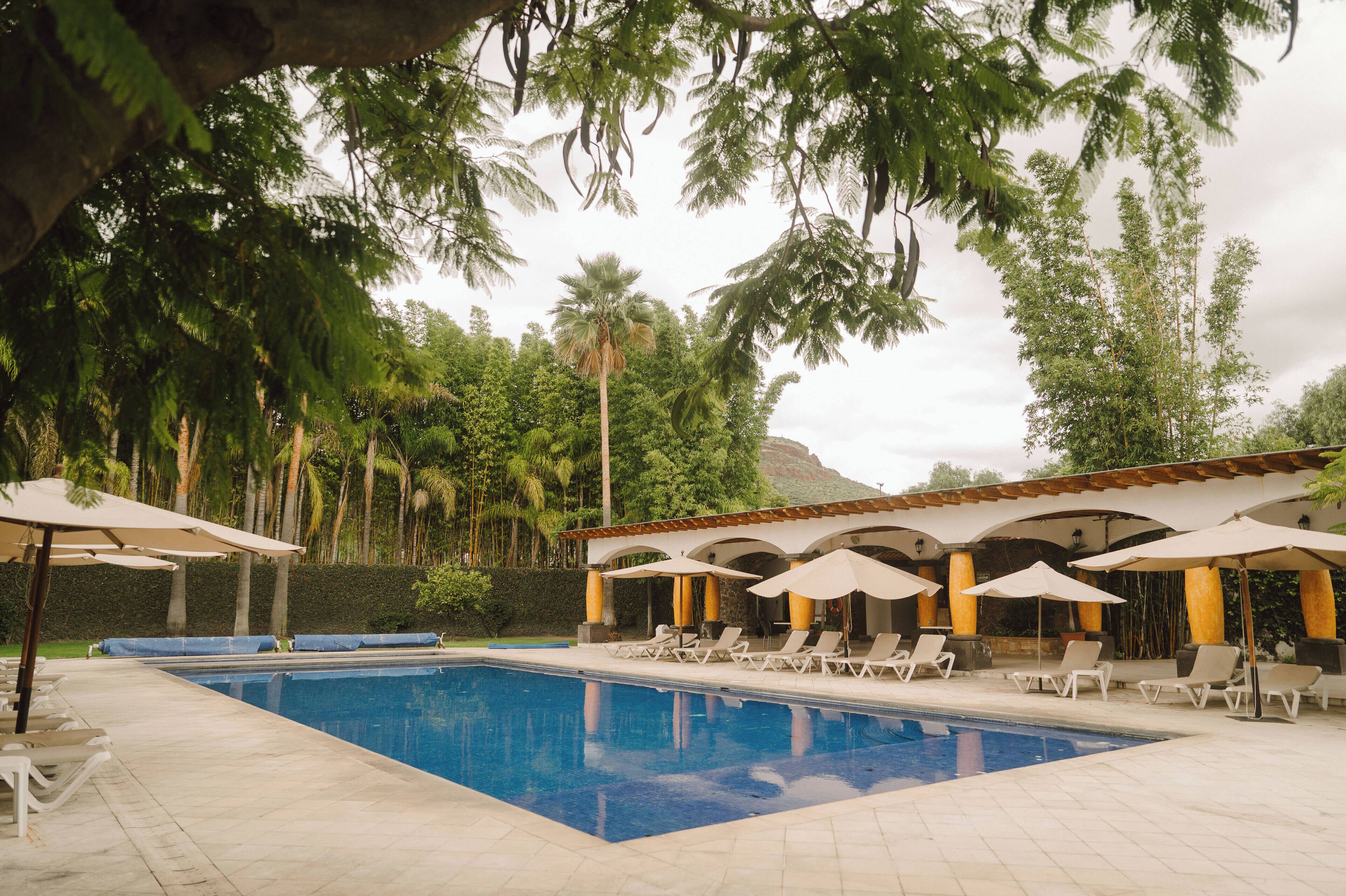 A pool area with lounge chairs and umbrellas, surrounded by palm trees and a building with arches.