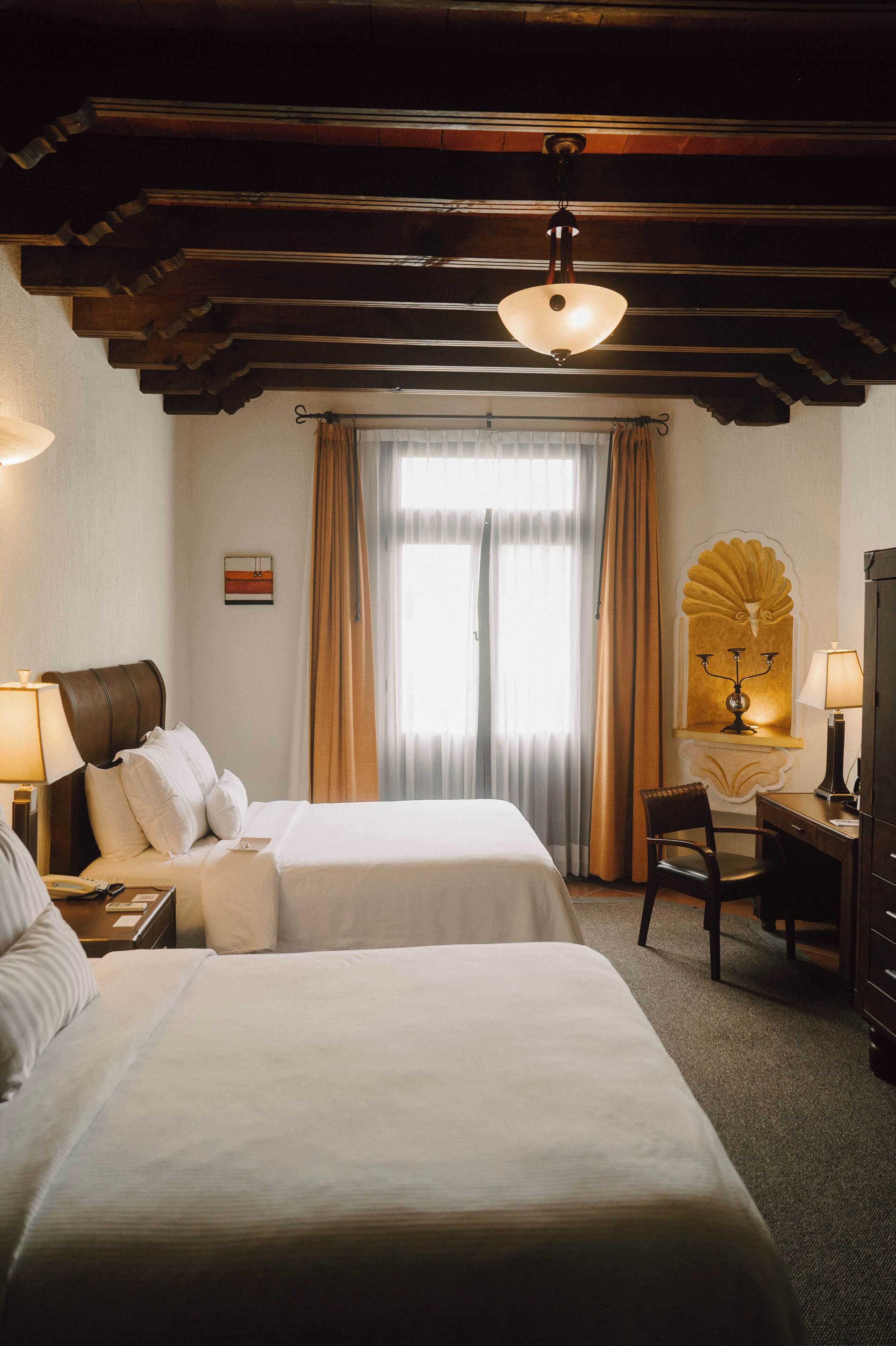 Habitación de hotel con dos camas, un escritorio, una silla, una lámpara y un ventanal con cortinas.