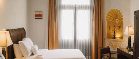 Habitación de hotel con dos camas, un escritorio, una silla, una lámpara y un ventanal con cortinas.