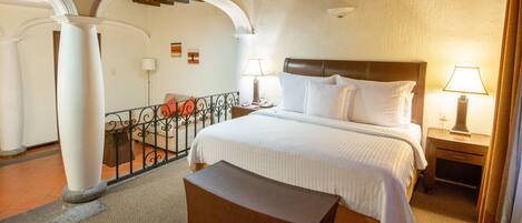 Habitación de hotel con una cama grande, vigas de madera en el techo, un balcón con barandilla de hierro forjado y un escritorio pequeño con una lámpara.