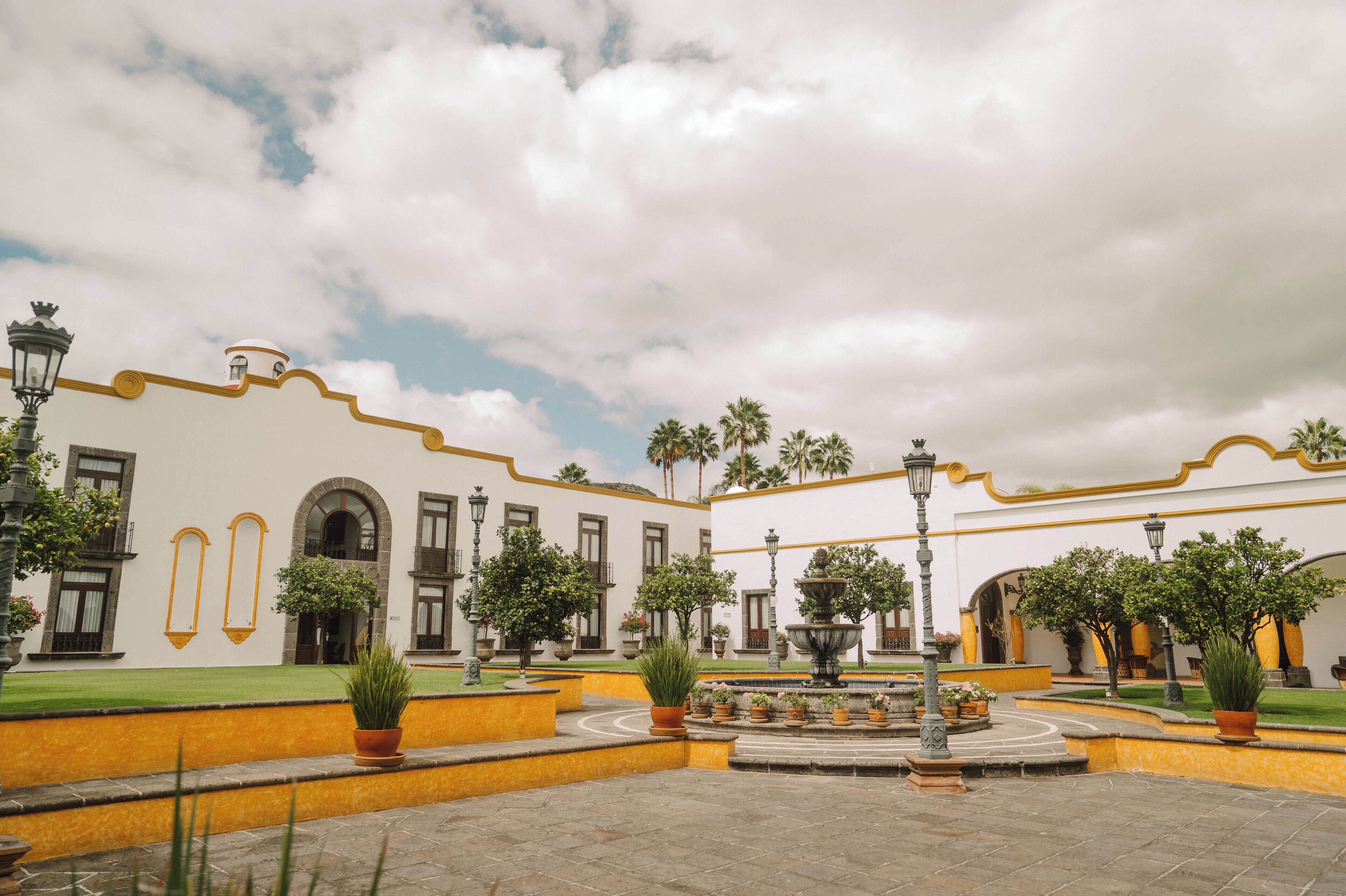A courtyard with a fountain, potted plants, and a building with arched windows and yellow trim.