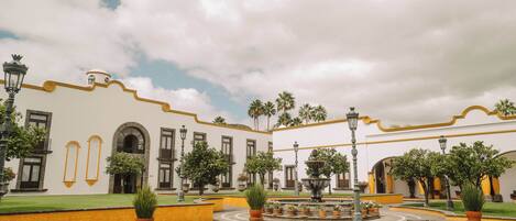A courtyard with a fountain, potted plants, and a building with arched windows and yellow trim.