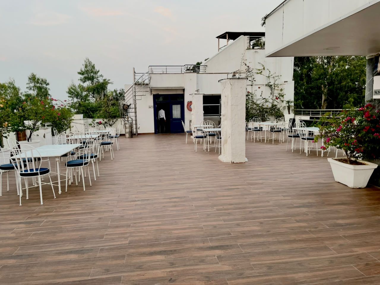 An outdoor dining area with white tables and chairs, a wooden floor, and a building entrance.