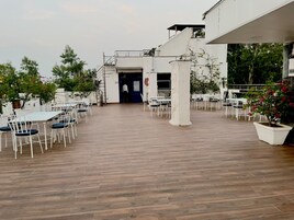 An outdoor dining area with white tables and chairs, a wooden floor, and a building entrance.