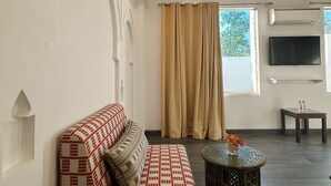 A living room with a patterned sofa, a small round table with flowers, a ceiling light fixture, and a large window with curtains. - Hotel Atulyaa Taj (Agra)