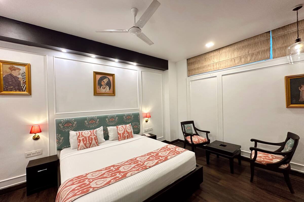 A hotel room with a bed, a desk, two chairs, a ceiling fan, and wall-mounted lights.