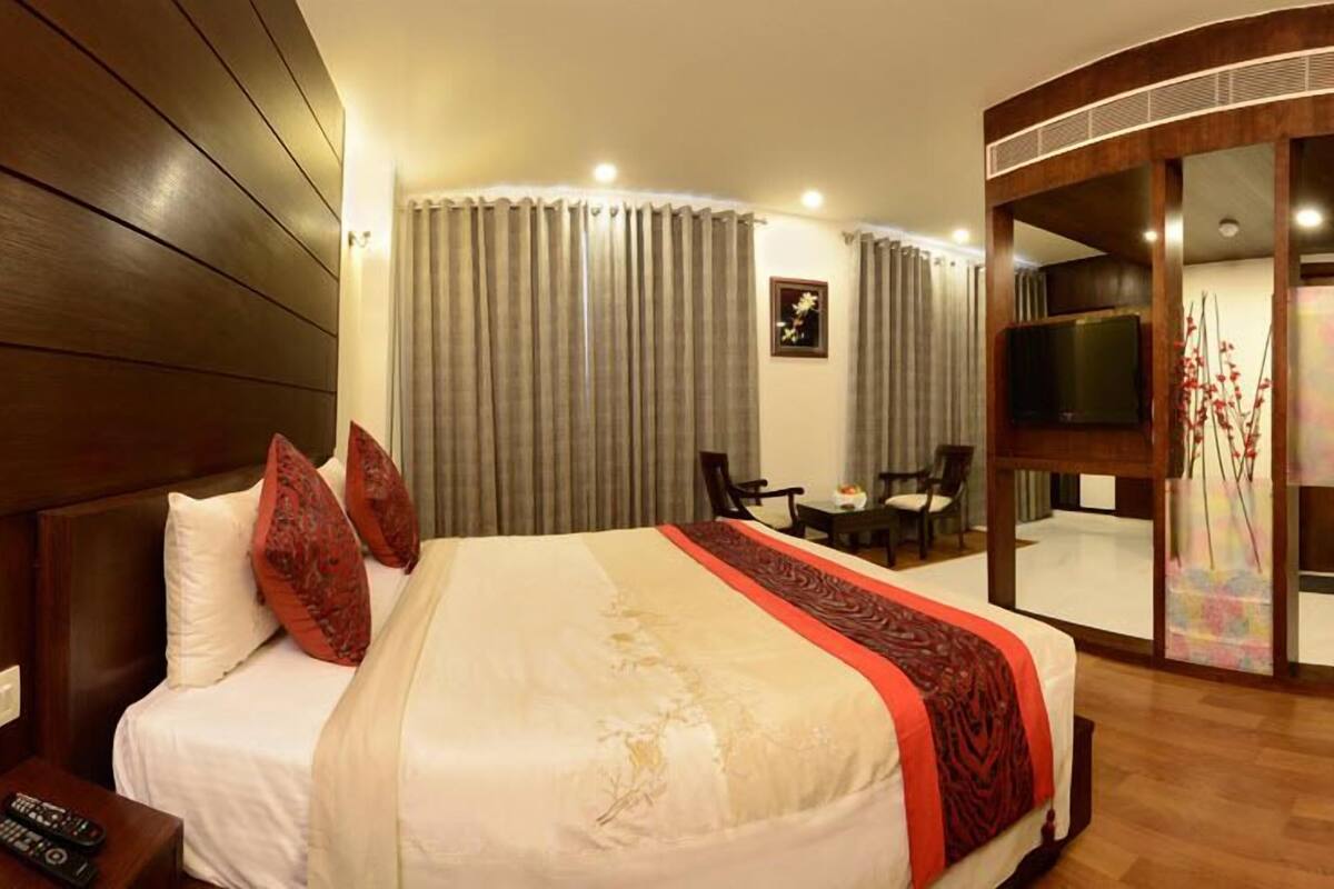 A hotel room with a bed, a TV, a desk with chairs, and a sliding door leading to a balcony.
