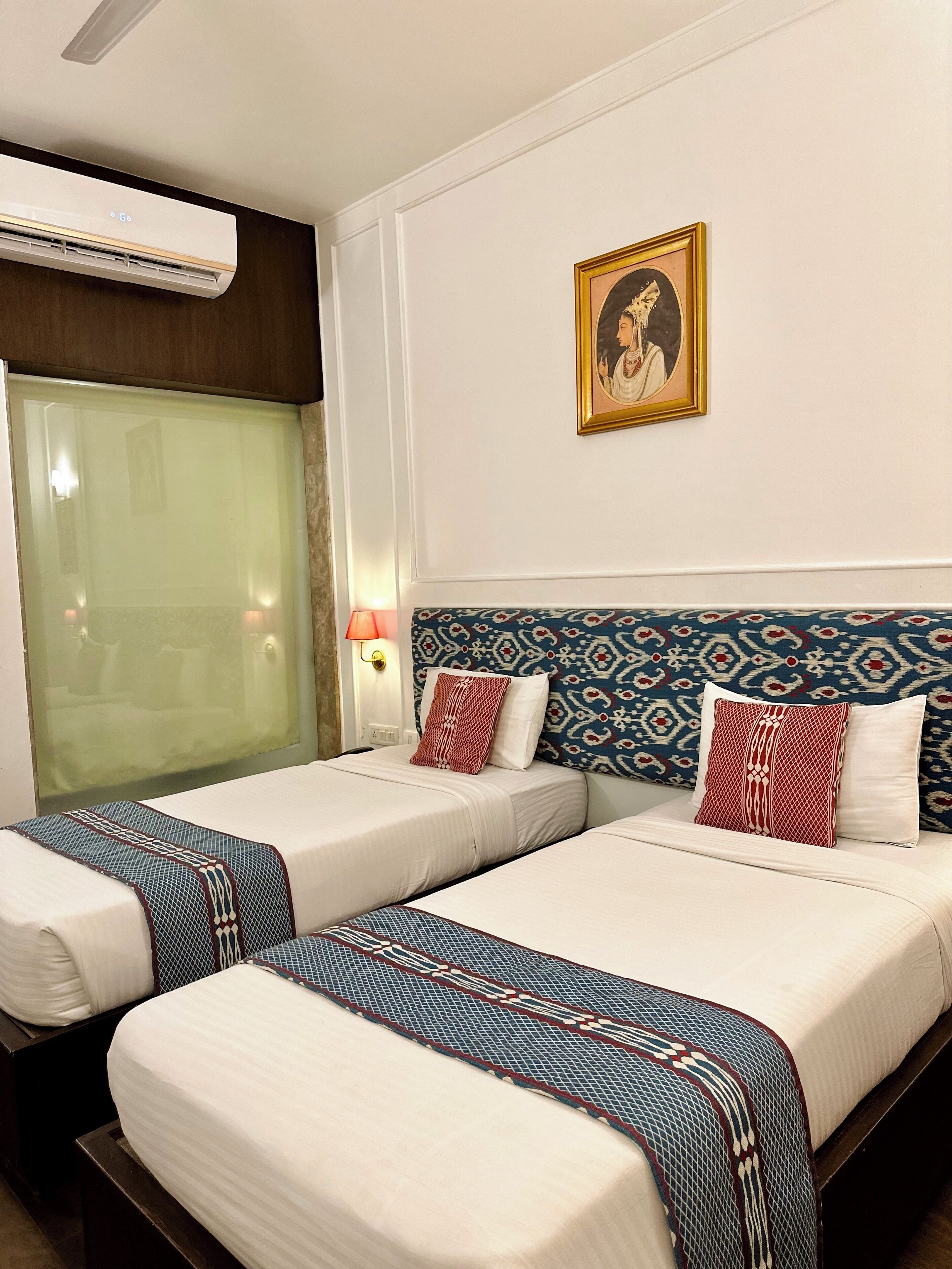 A hotel room with two beds, a large window with a view, an air conditioning unit, a wall-mounted lamp, and a framed picture.