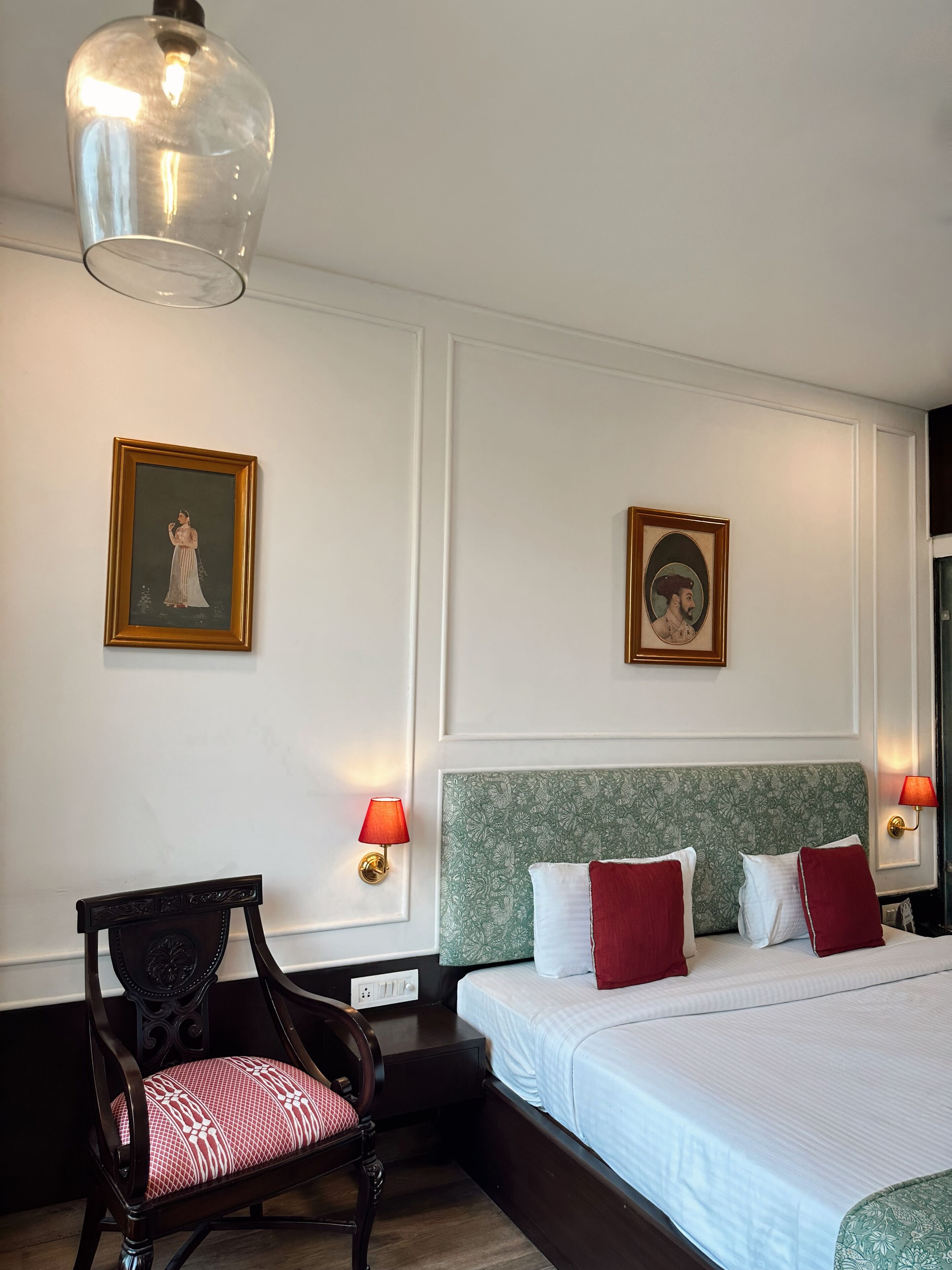 Hotellihuone, jossa on sänky, tuoli, kaksi seinälle asennettua lamppua ja kaksi kehystettyä kuvaa.