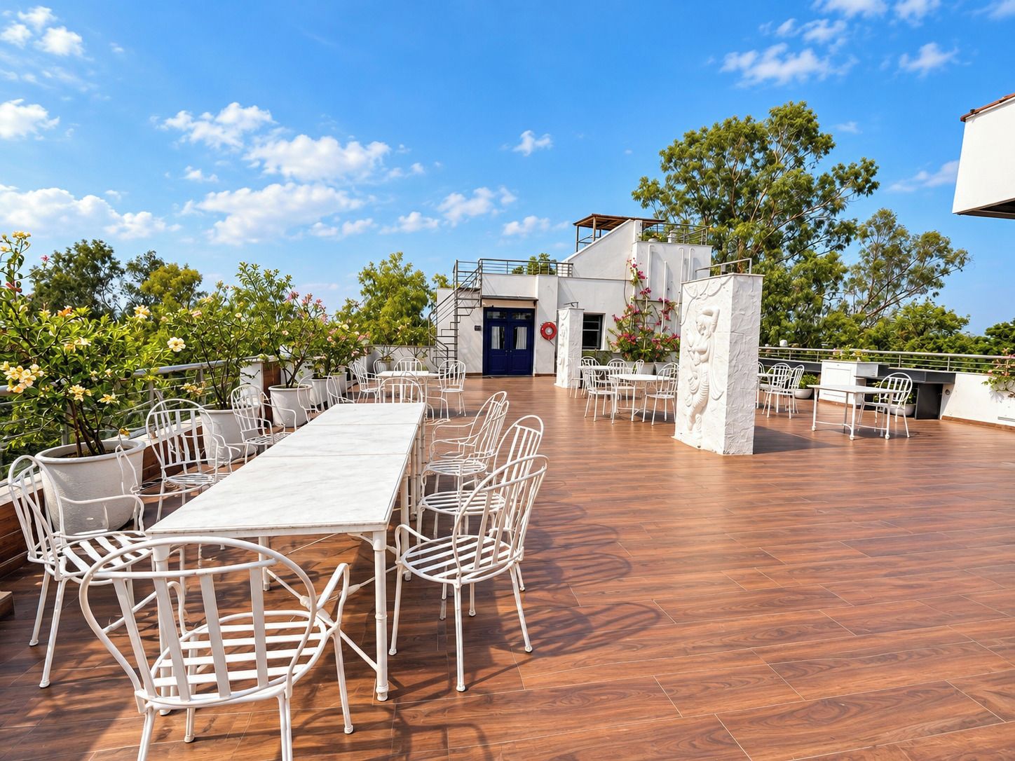 An outdoor dining area with white metal chairs and tables, surrounded by potted plants and trees.