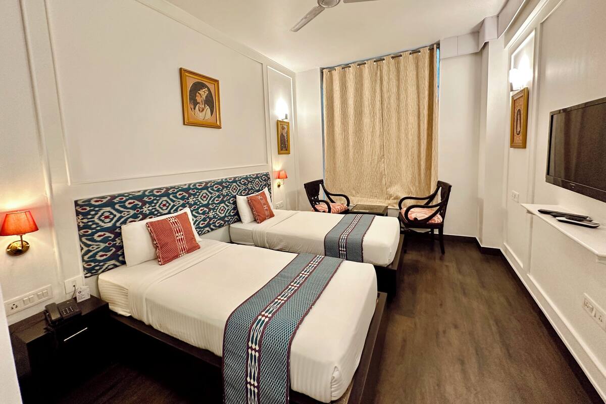 A hotel room with two beds, a desk, a chair, a TV, and a ceiling fan.