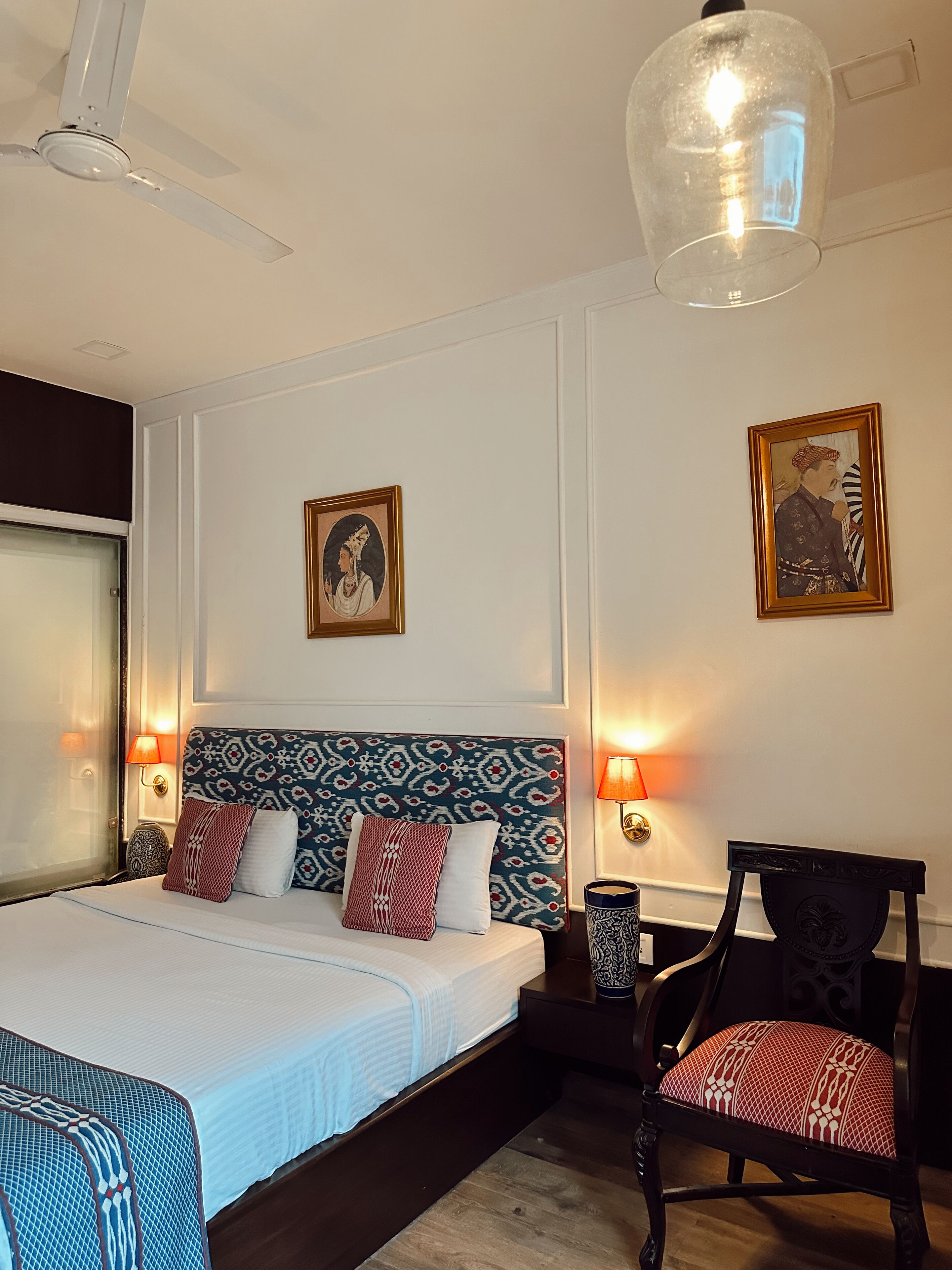 A hotel room with a large bed, two wall-mounted lamps, a ceiling fan, and two framed pictures on the walls.
