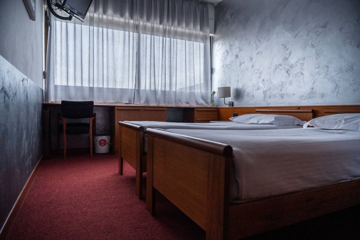 Une chambre d’hôtel avec un lit en bois, un bureau avec une chaise, une fenêtre avec des rideaux légers et un mur orné d’un motif texturé.