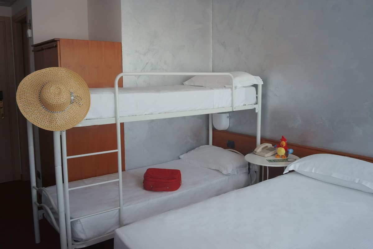 Une chambre avec un lit superposé, un chapeau de paille posé sur le lit du haut, une valise rouge sur le lit du bas, et une petite table avec un téléphone et un coq en jouet.