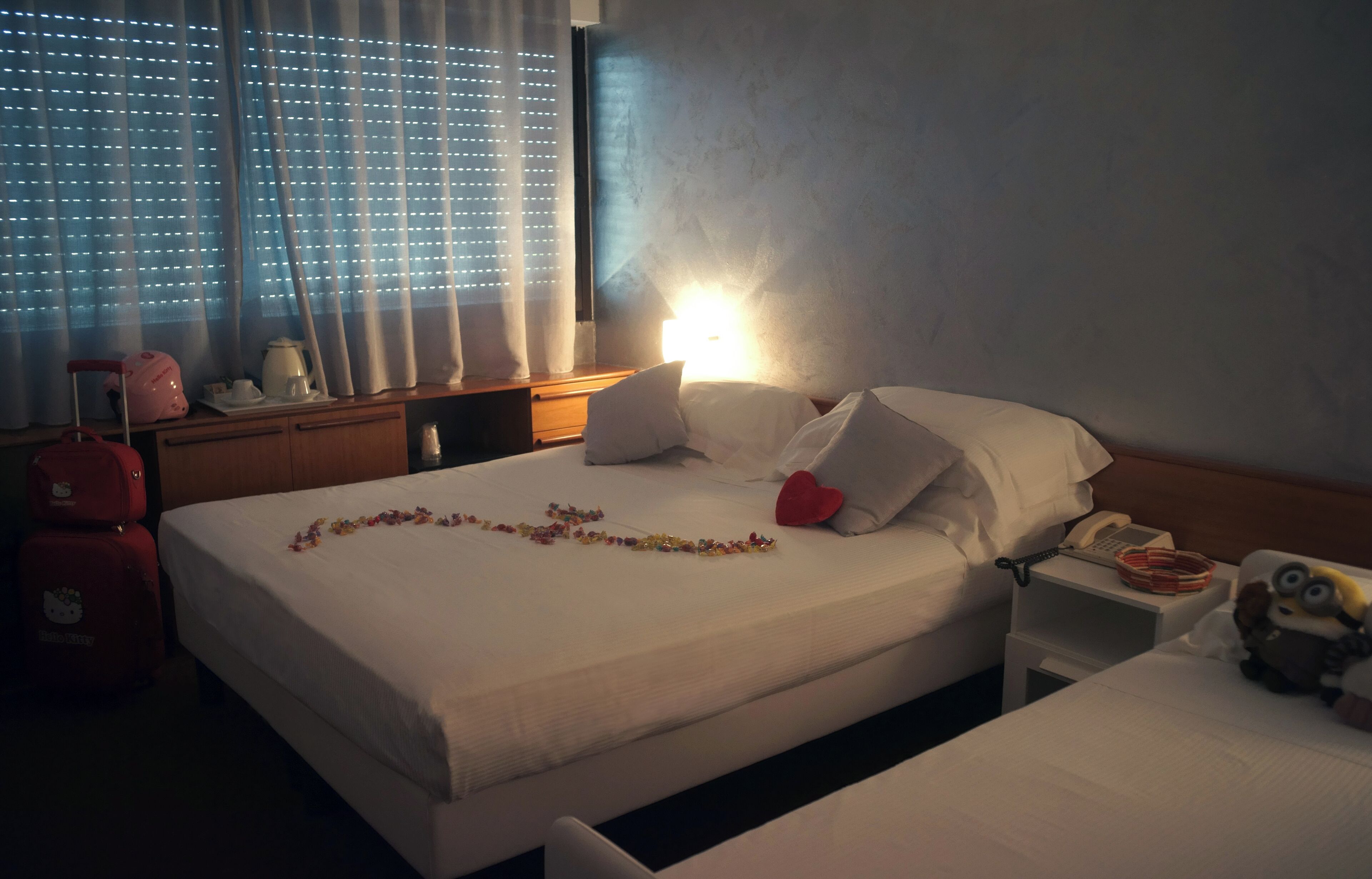 A hotel room with a bed, bedside table, and a window with blinds.