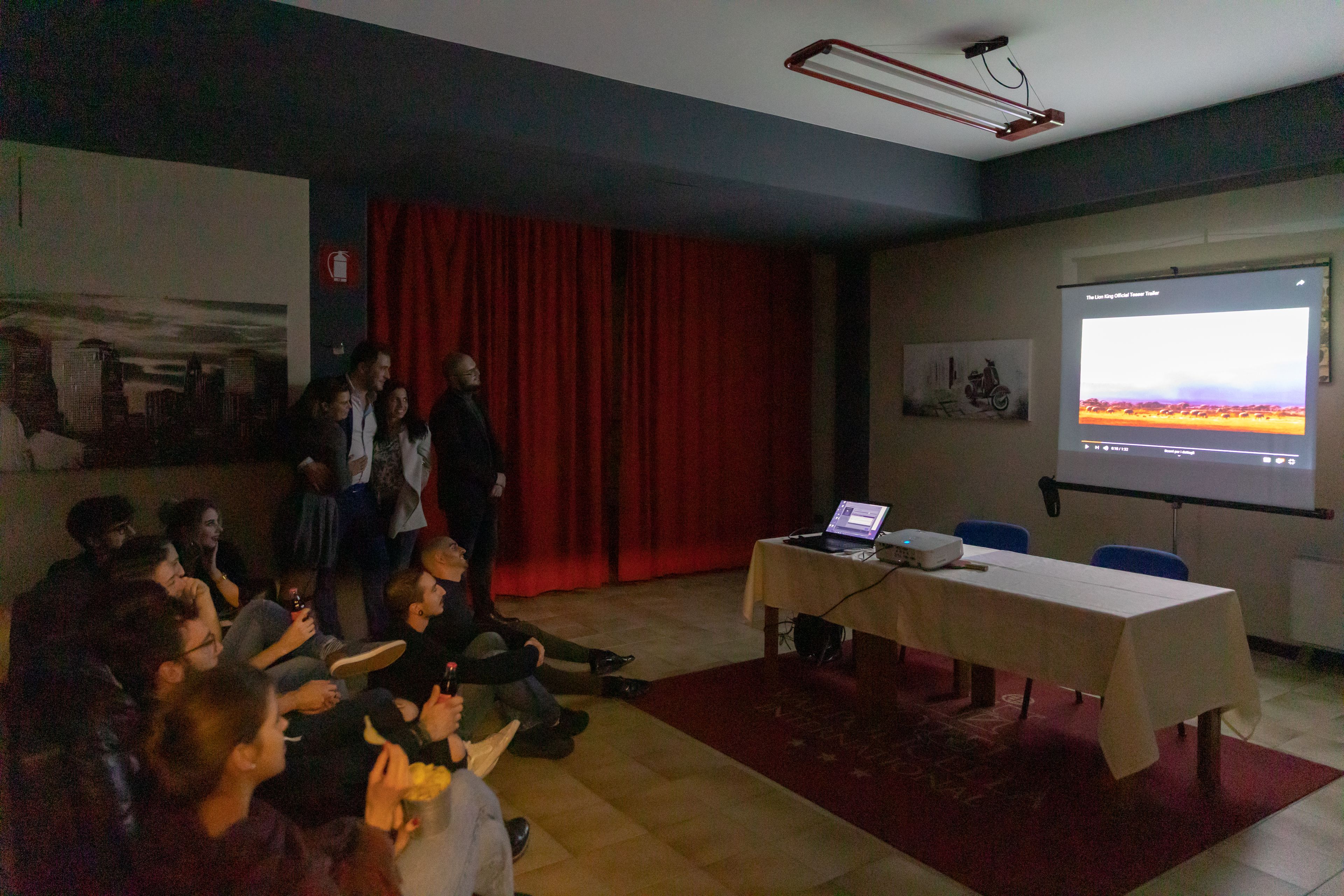 A conference room with a projector screen, a table with a laptop, and attendees seated.