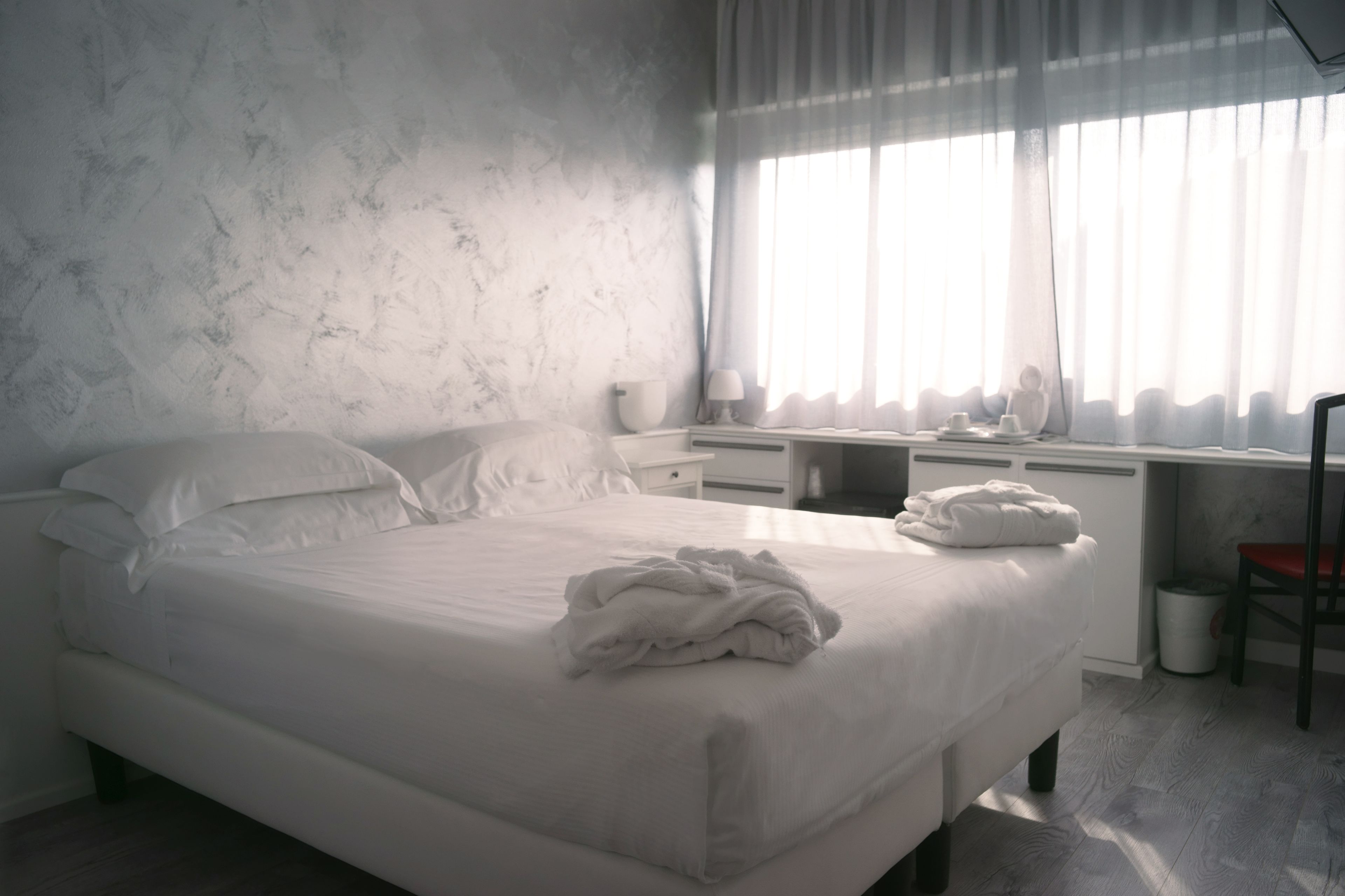 A neatly made bed with a white comforter and pillows, a bedside table with a lamp, a chair, and a desk with a chair.