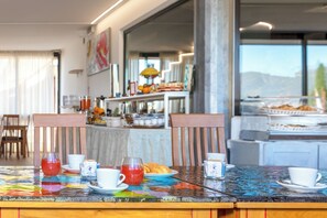 Une table de petit-déjeuner avec des tasses et un pichet, un buffet rempli de nourriture, et une vue sur l’extérieur.
