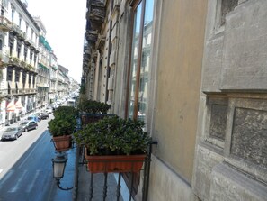 A narrow street with buildings on either side, potted plants on a balcony, and a street lamp.