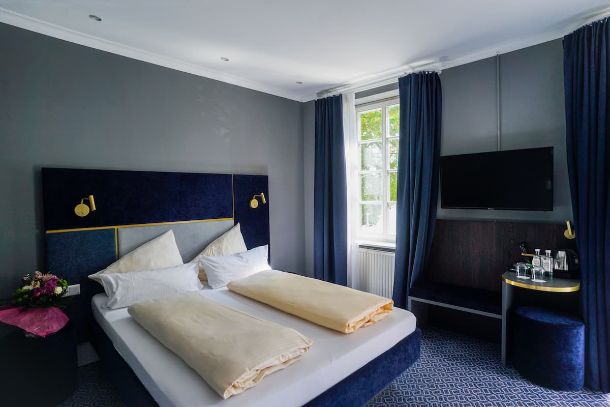 A hotel room with a large bed, a flat-screen TV, a small table with toiletries, and a window with curtains.