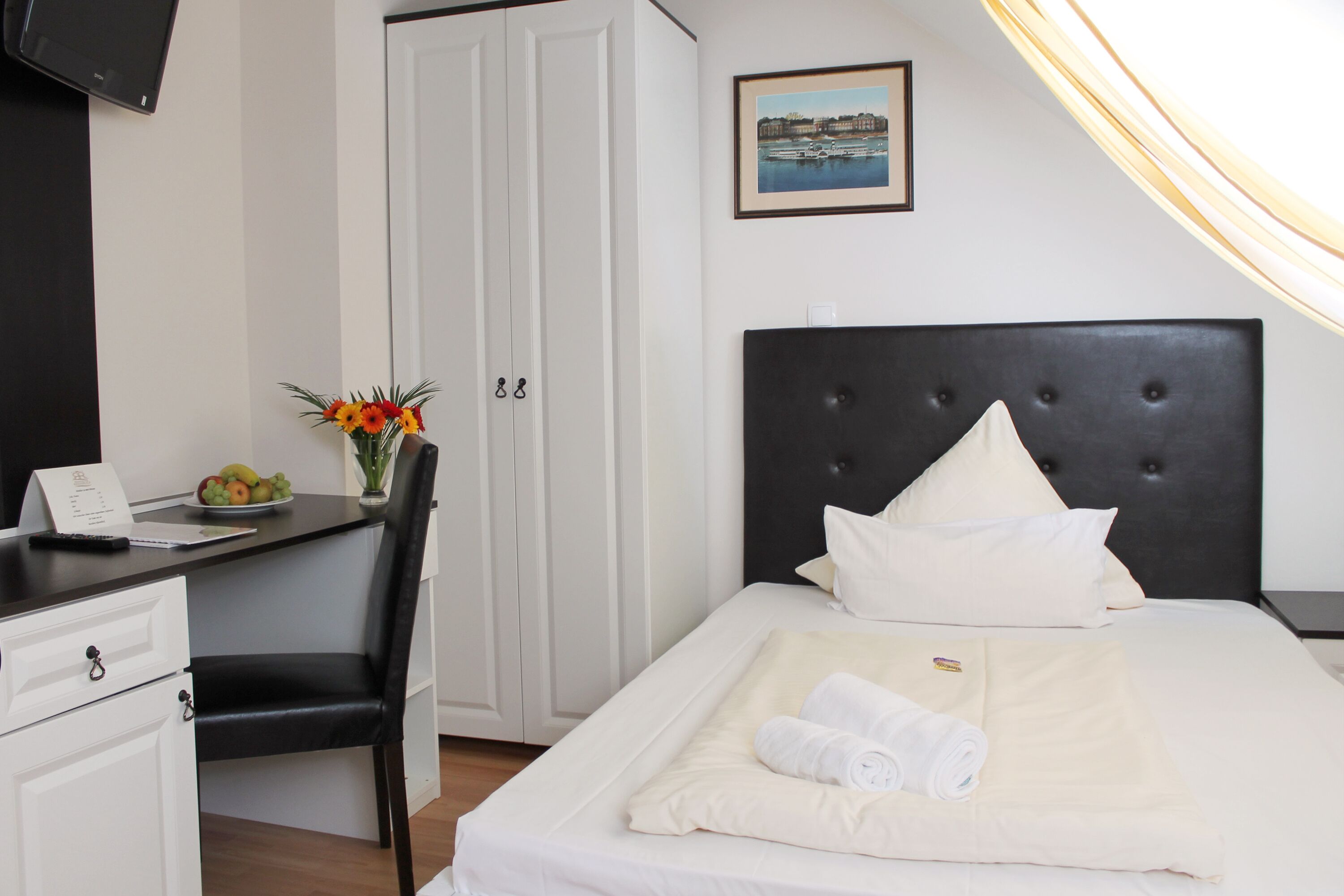 Een hotelkamer met een bed, een bureau met een stoel, een tafeltje met fruit, een vaas met bloemen en een schilderij aan de muur.