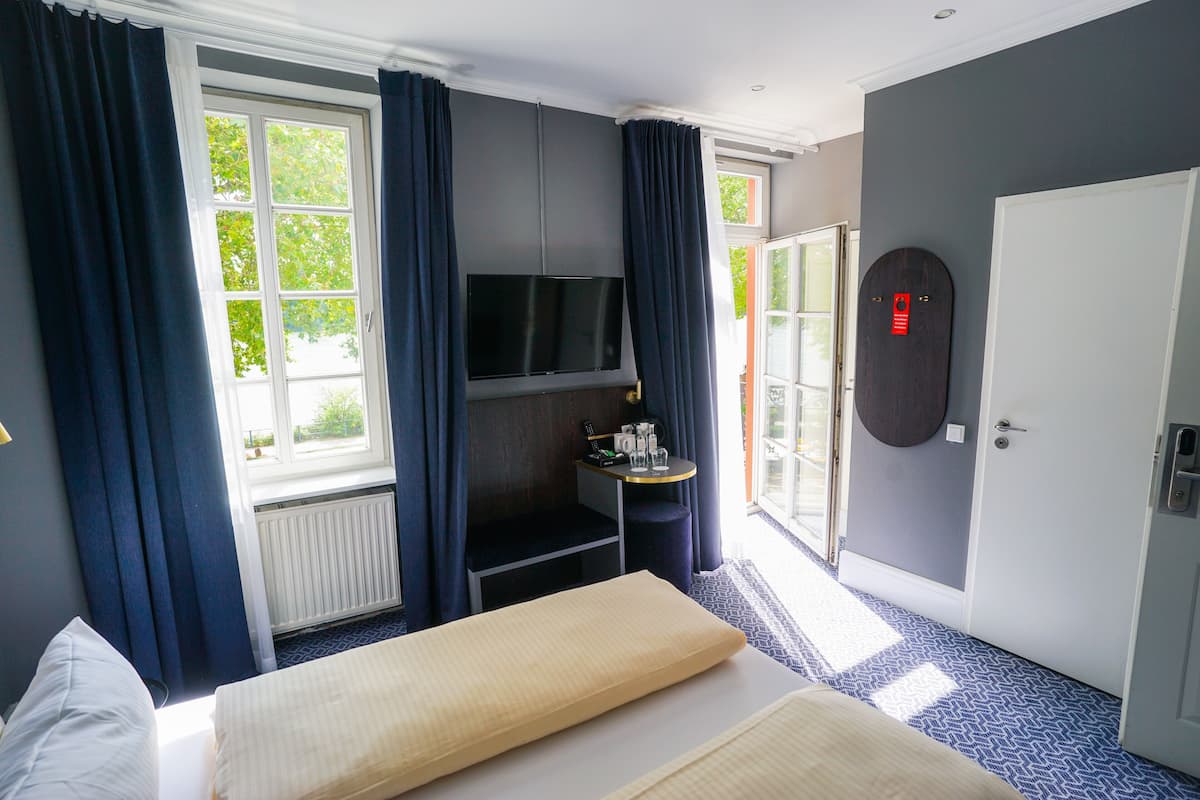 A hotel room with a bed, a TV mounted on the wall, a small table with a bottle and glasses, and a door with a red lock.