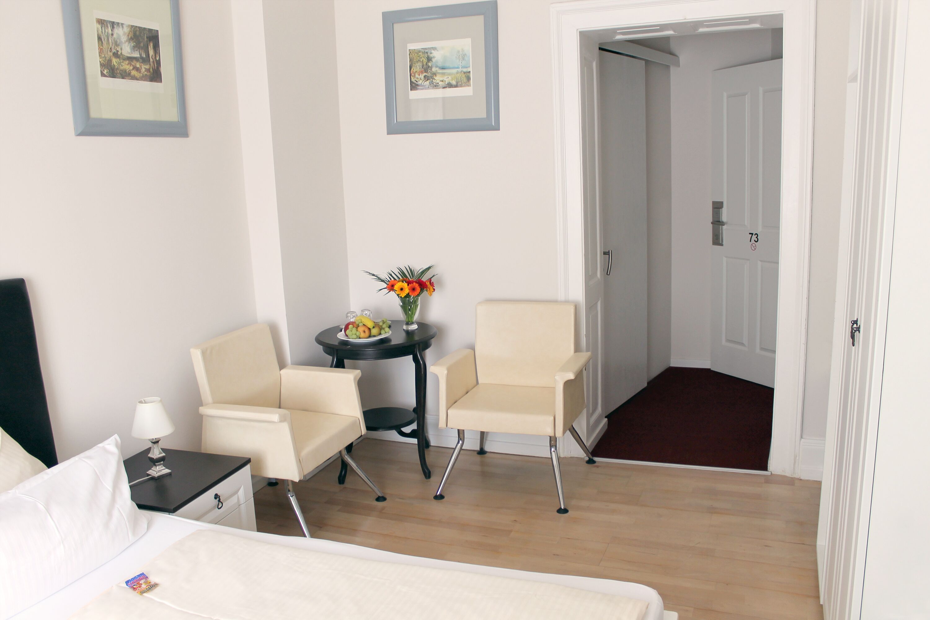 A hotel room with a bed, two chairs, a small table with fruit and flowers, and a door marked with the number 73.