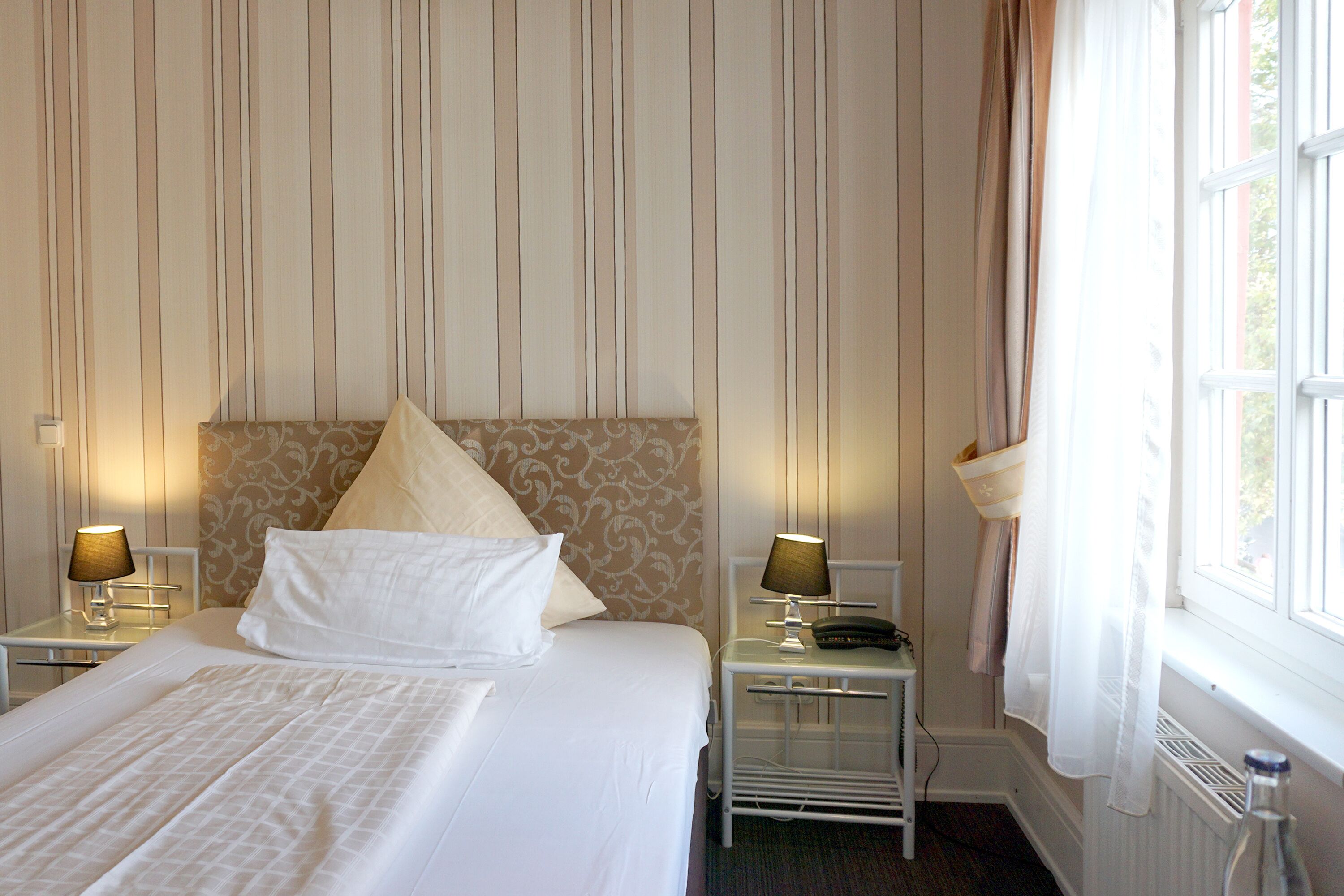 A hotel room with a large bed, two bedside tables with lamps, a nightstand, and a window with curtains.