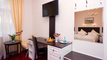 A hotel room with a desk, chair, TV, bed, and a small table with fruits and flowers.