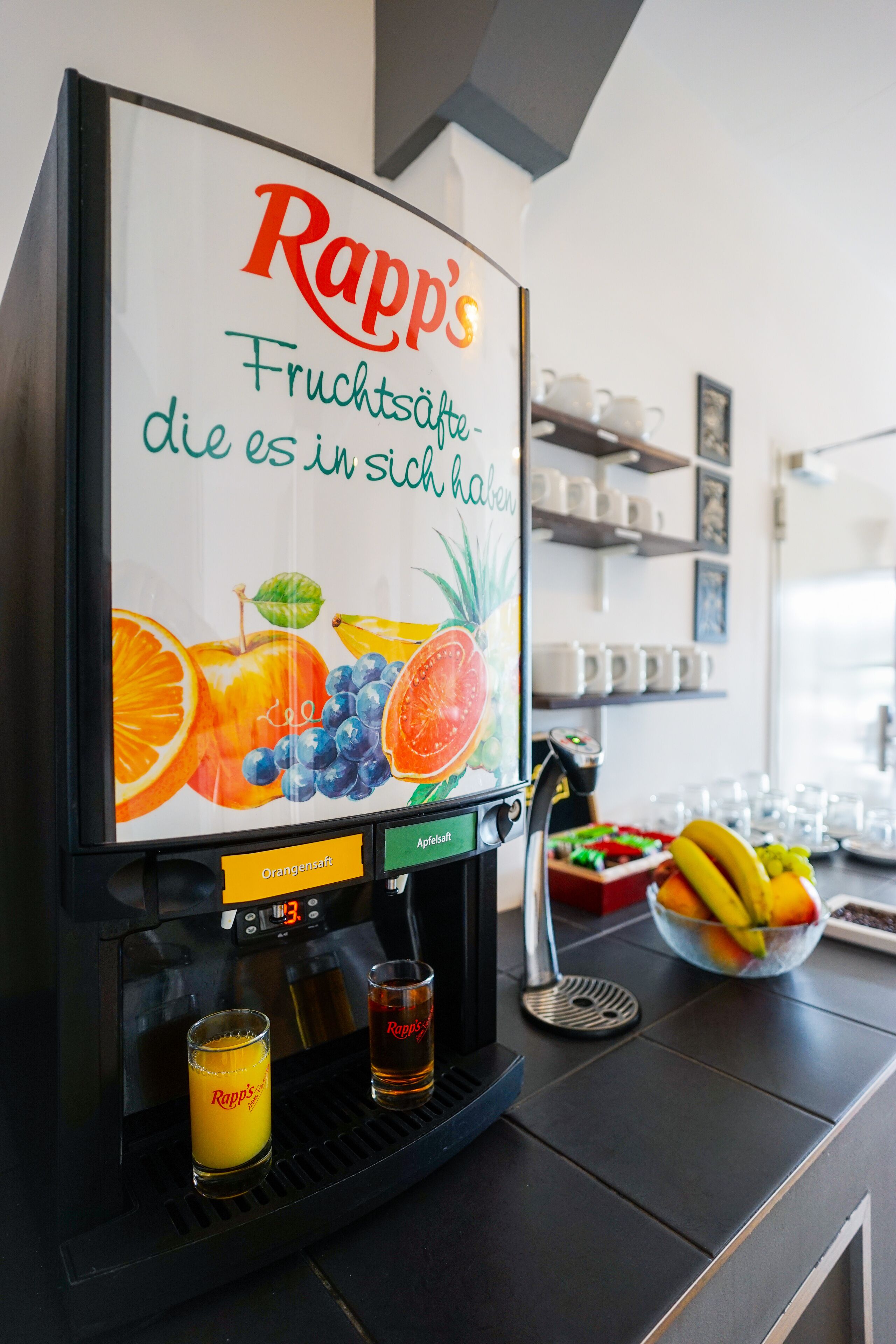 A coffee machine with a Rapp's fruit juice dispenser, a glass of orange juice, and a glass of Rapp's juice.