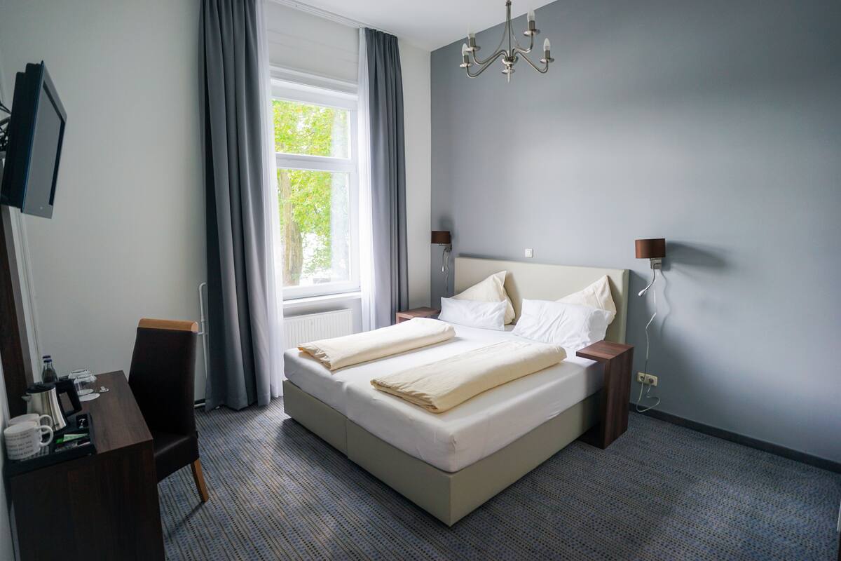 A hotel room with a bed, bedside tables, a desk with a chair, a TV mounted on the wall, and a window with curtains.