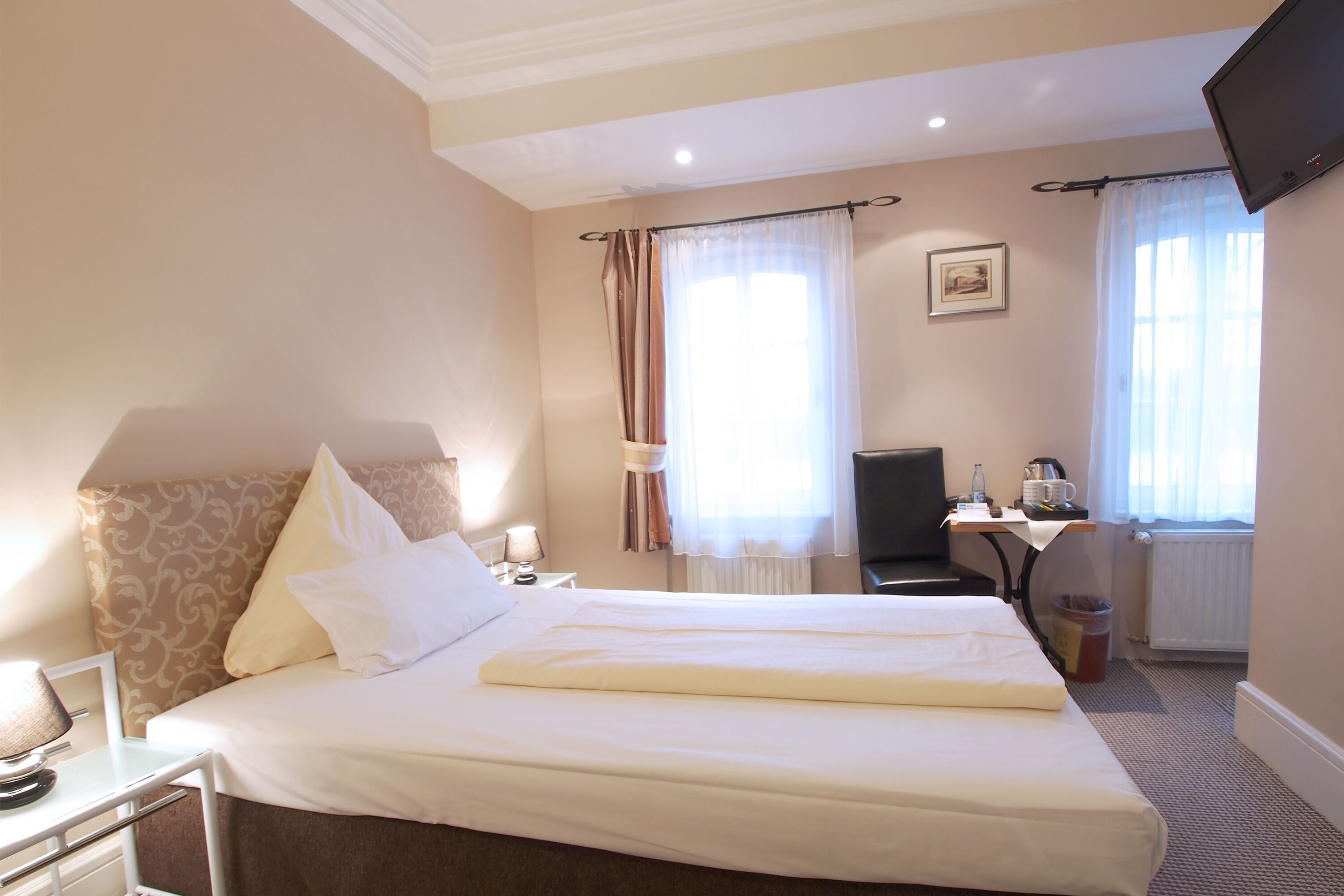 Habitación de hotel con una cama grande, un escritorio con silla, televisor montado en la pared y una ventana con cortinas.