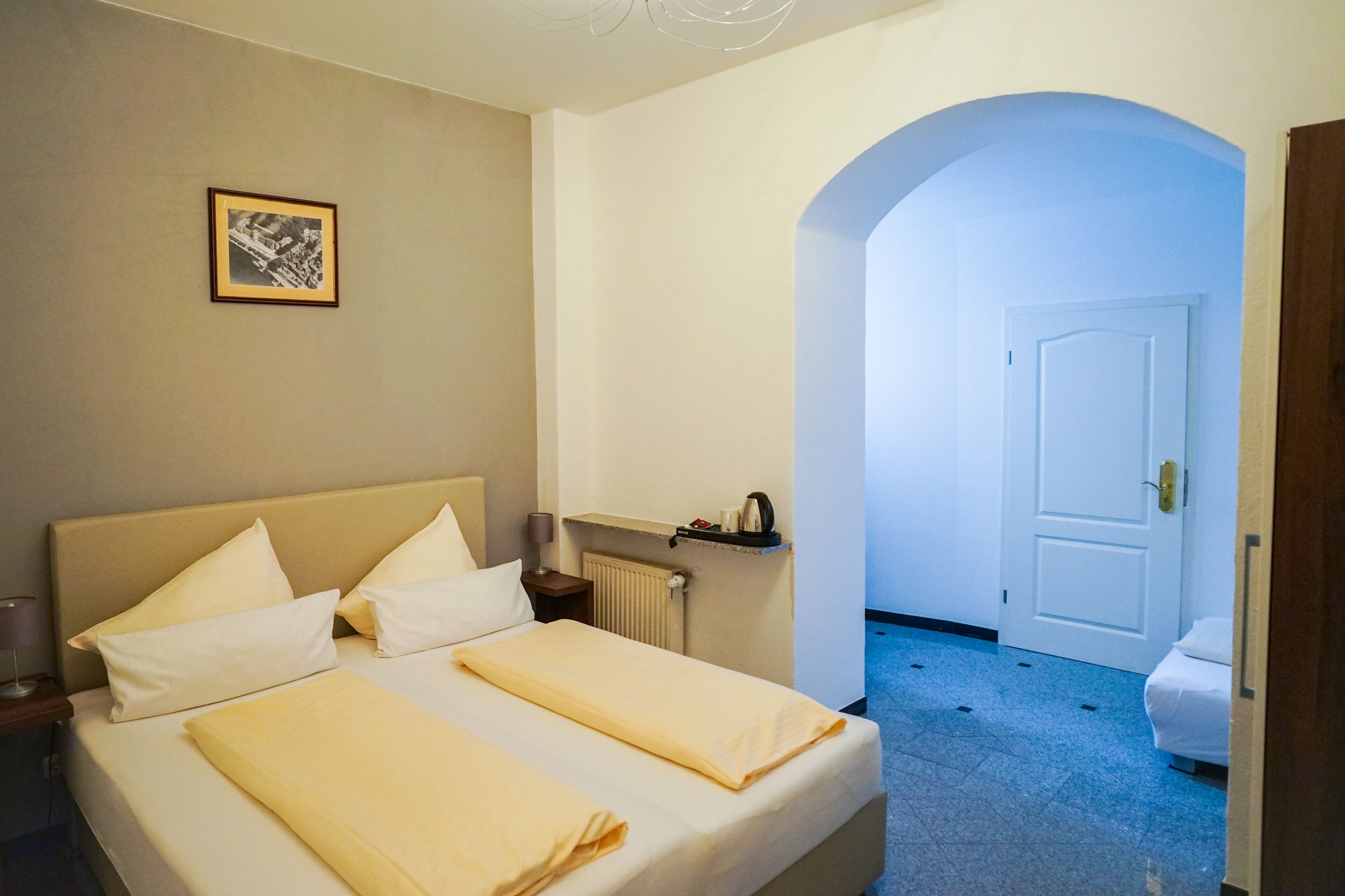 Habitación de hotel con cama, mesita de noche, un cuadro en la pared y una puerta que da a otra habitación.