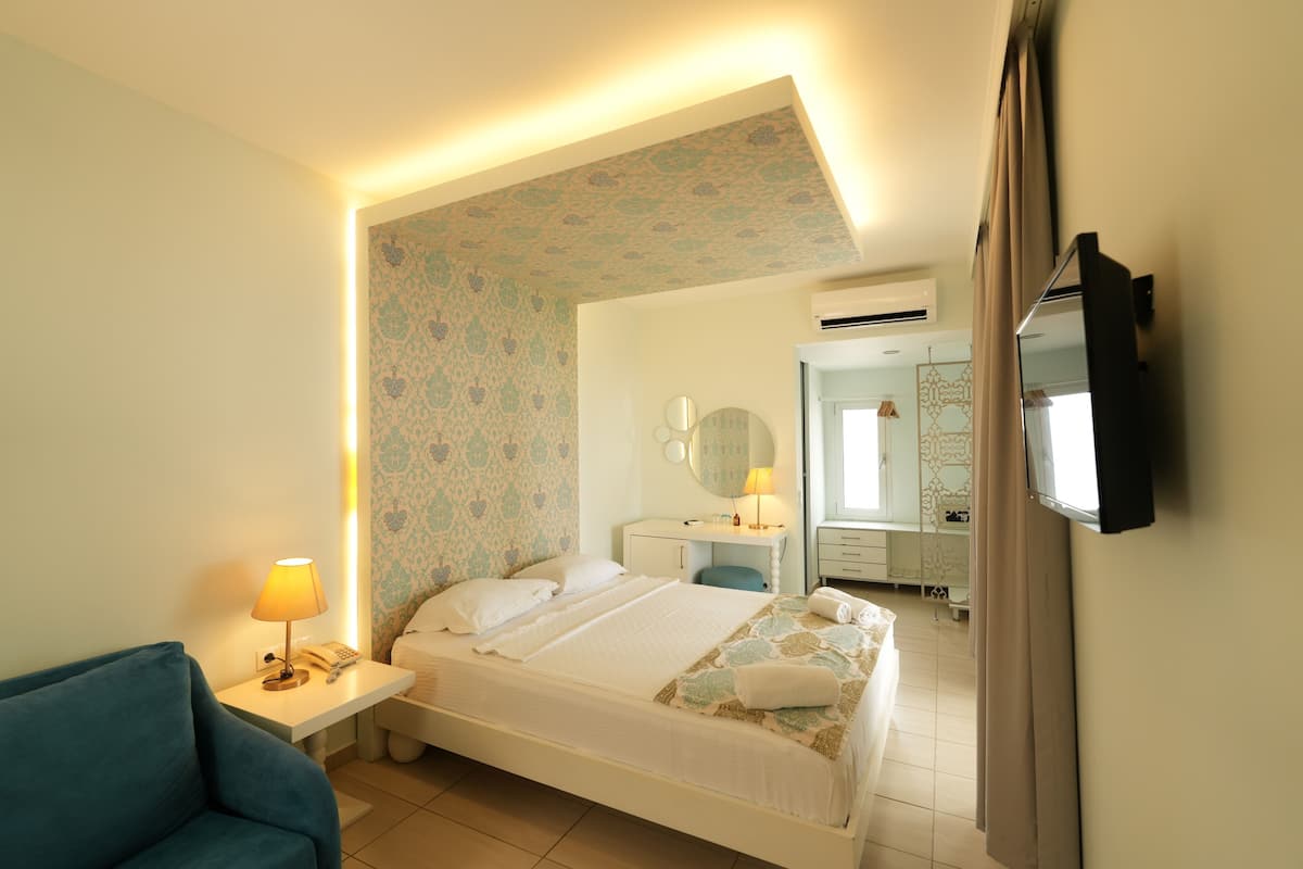 A hotel room with a bed, bedside table with a lamp, a TV mounted on the wall, and a blue armchair.