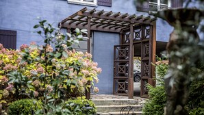 En pergola av trä med ett geometriskt mönster, en stenlagd gång som leder till en dörr och frodigt grönska, inklusive rosa trädgårdshortensior.