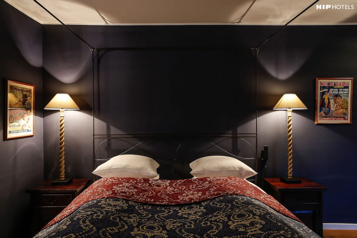 A hotel room with a dark blue wall, a bed with a red and blue patterned bedspread, two bedside lamps, and a poster on the wall.