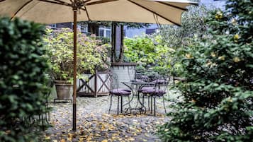 Un patio al aire libre con mesa y sillas bajo un paraguas grande, rodeado de plantas en macetas y un suelo de baldosas de piedra.