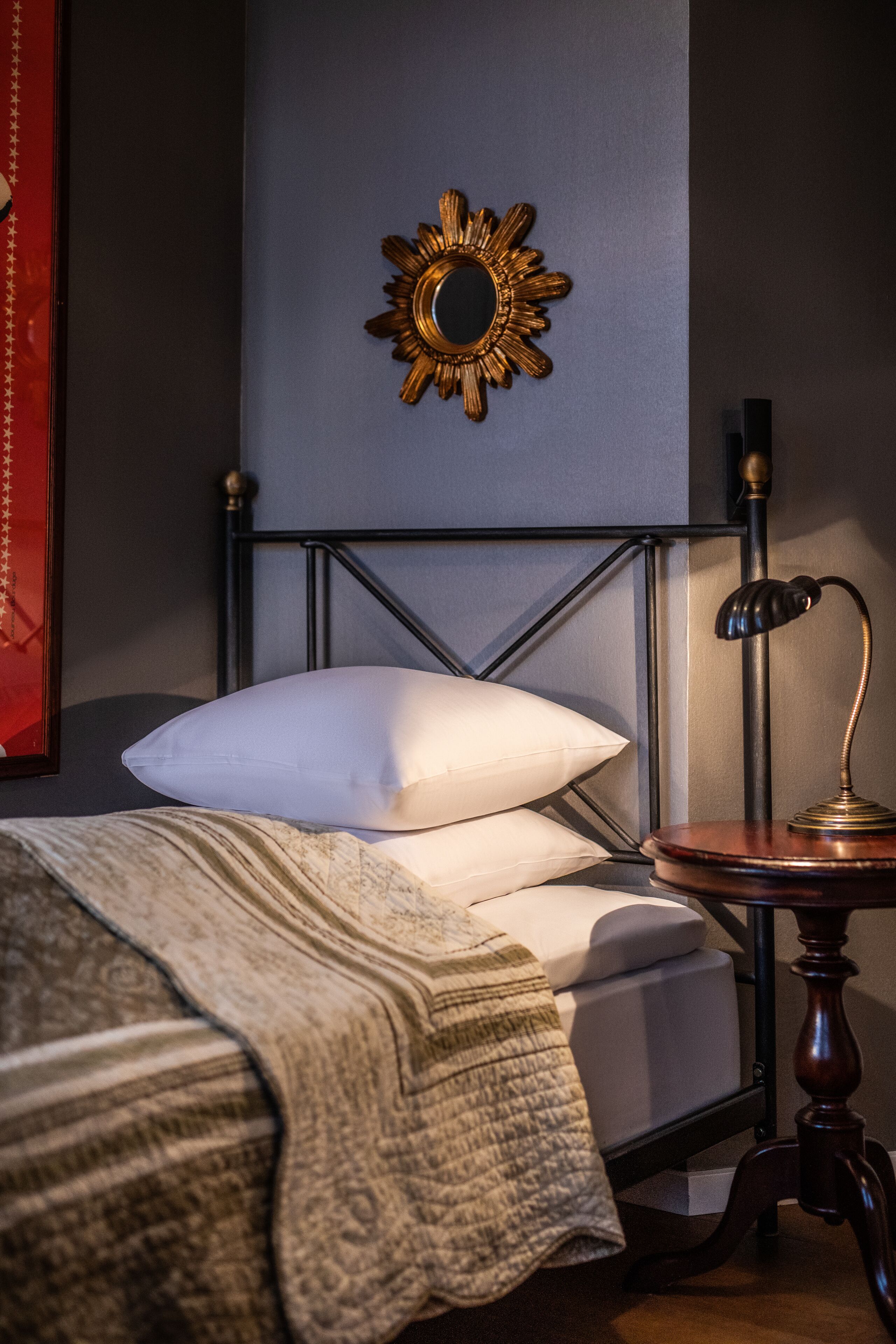 A neatly made bed with a white pillow and a patterned blanket, a round table with a lamp, and a sunburst mirror on the wall.
