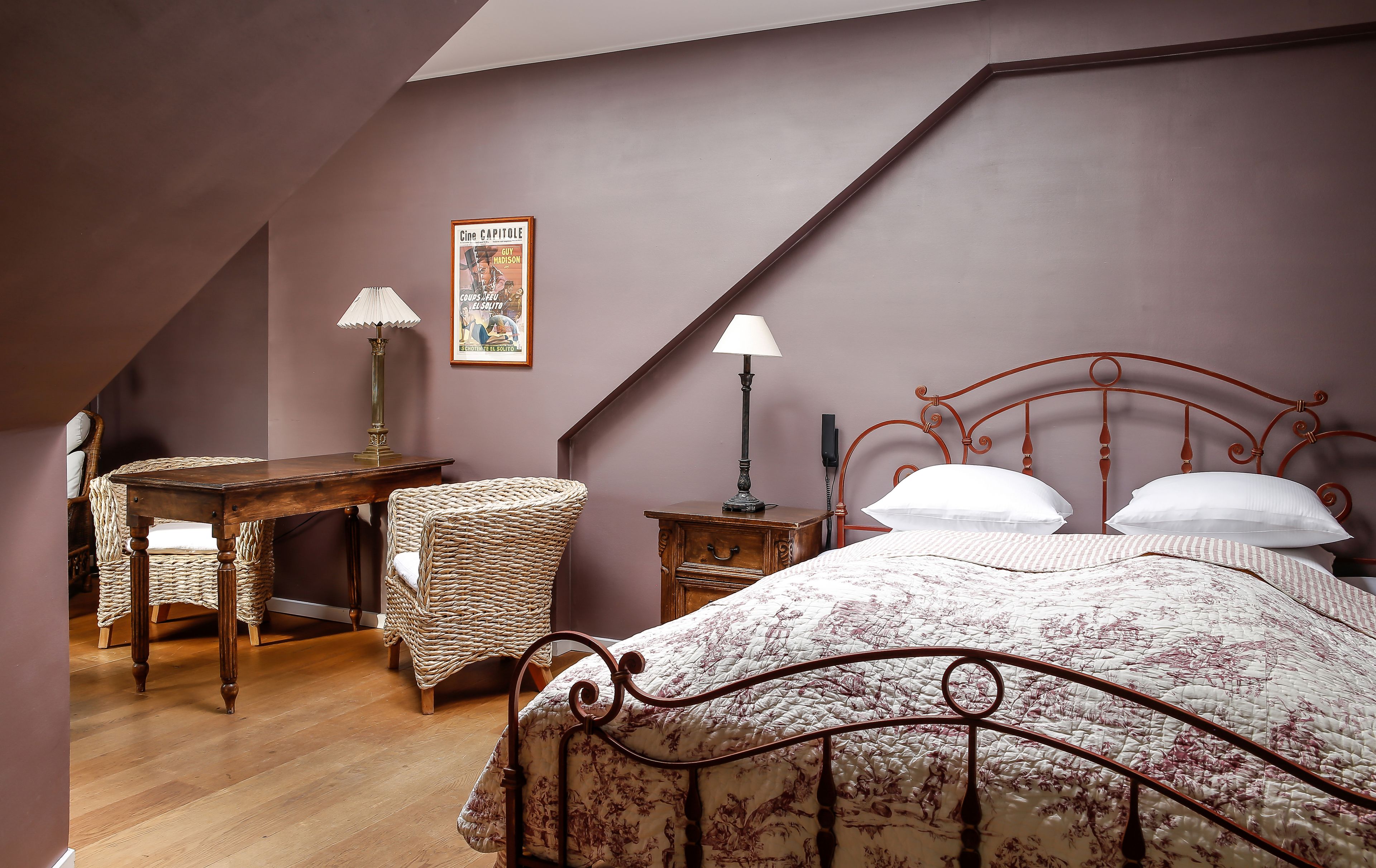 Un dormitorio con techo inclinado, un escritorio de madera con lámpara, una silla de mimbre, una cama con colcha estampada y un cartel en la pared.